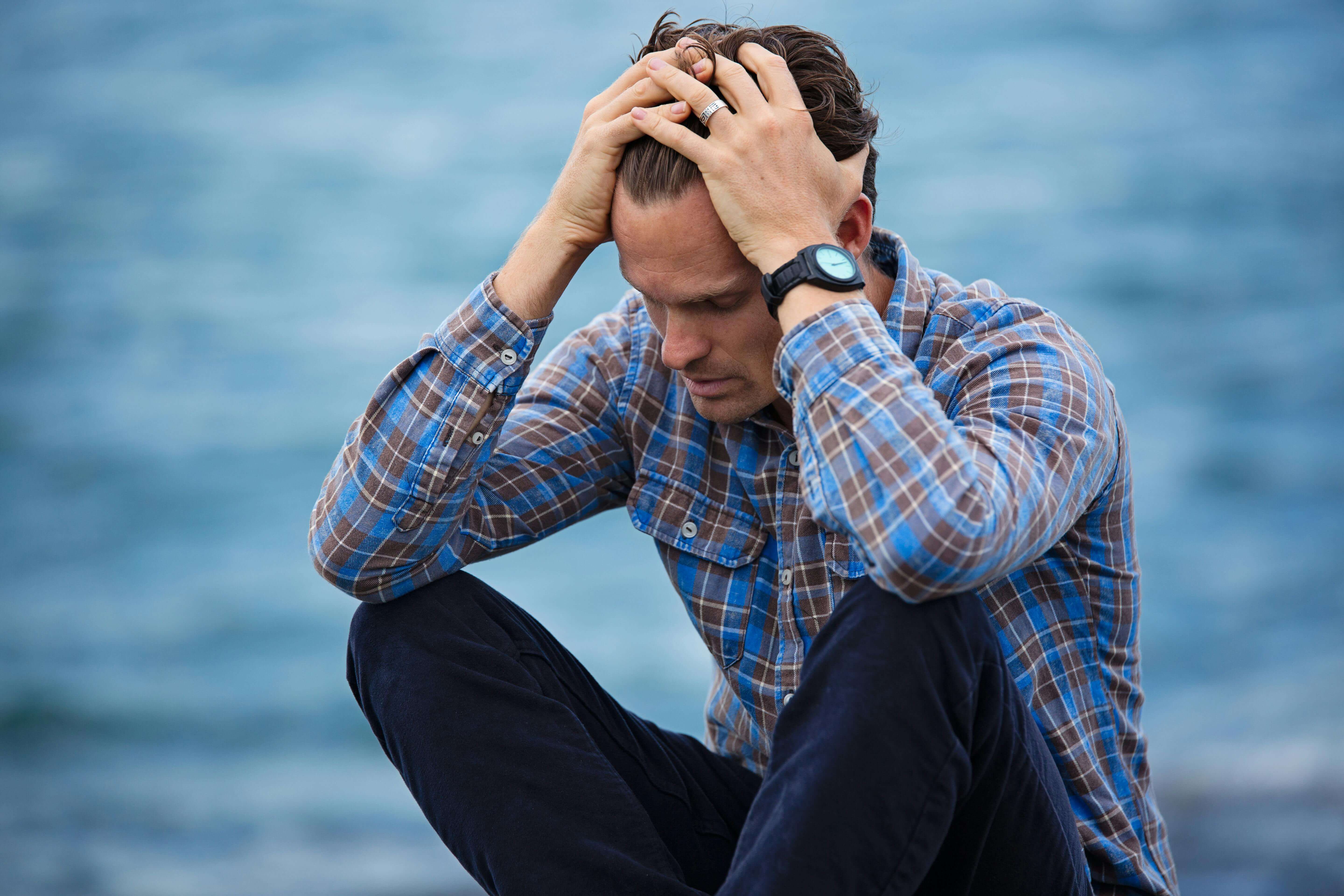 Upset man sitting on the ground in front of water running his hands through his hair. Heal from your anxiety symptoms and reclaim your life with the support of a men's therapist in Rancine, WI.