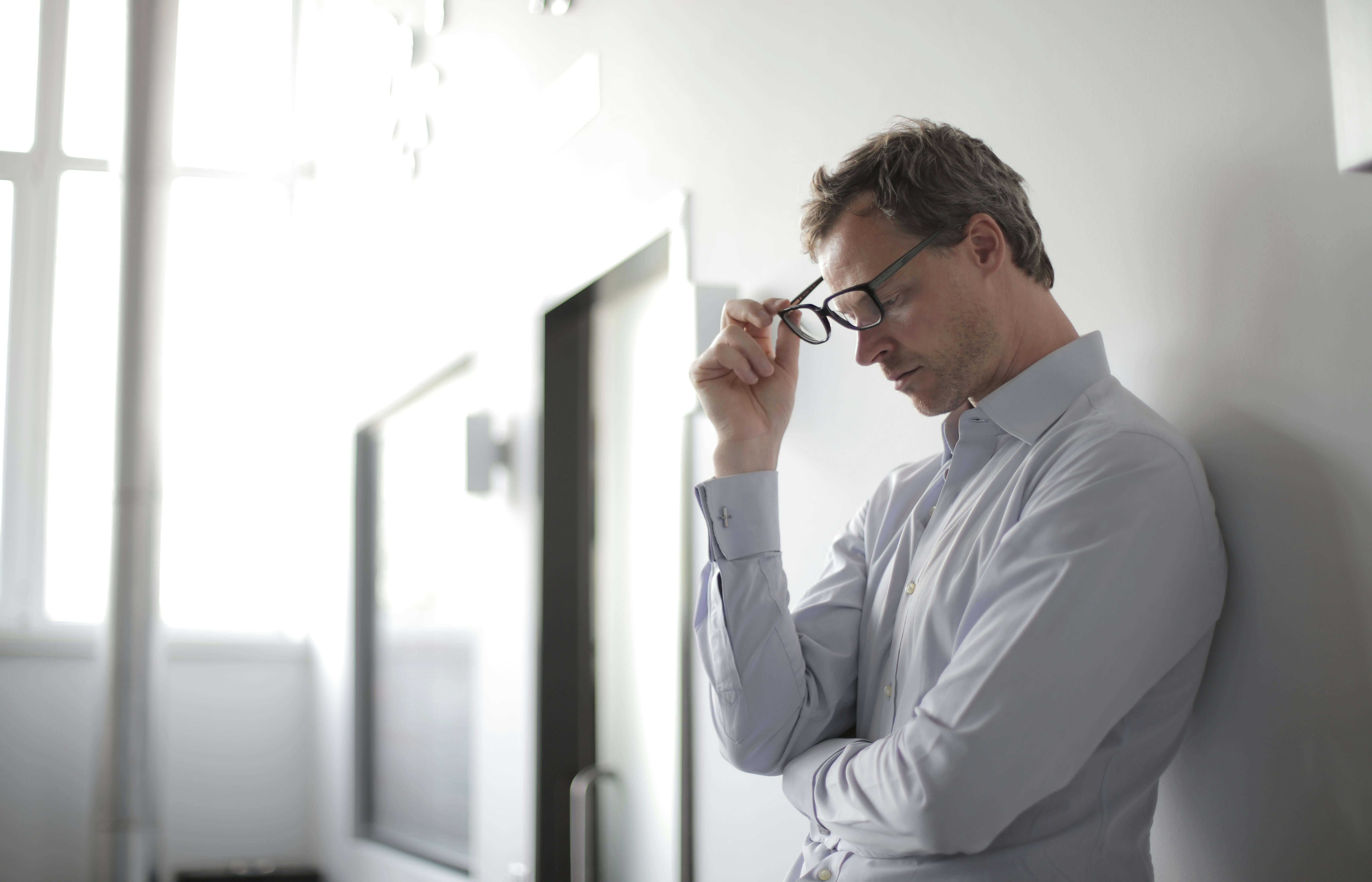 A thoughtful businessman stands against a wall, holding his glasses with a pained expression. Find clarity and calm through men’s therapy in Kenosha, WI helping high-performance men overcome anxiety and reclaim balance.