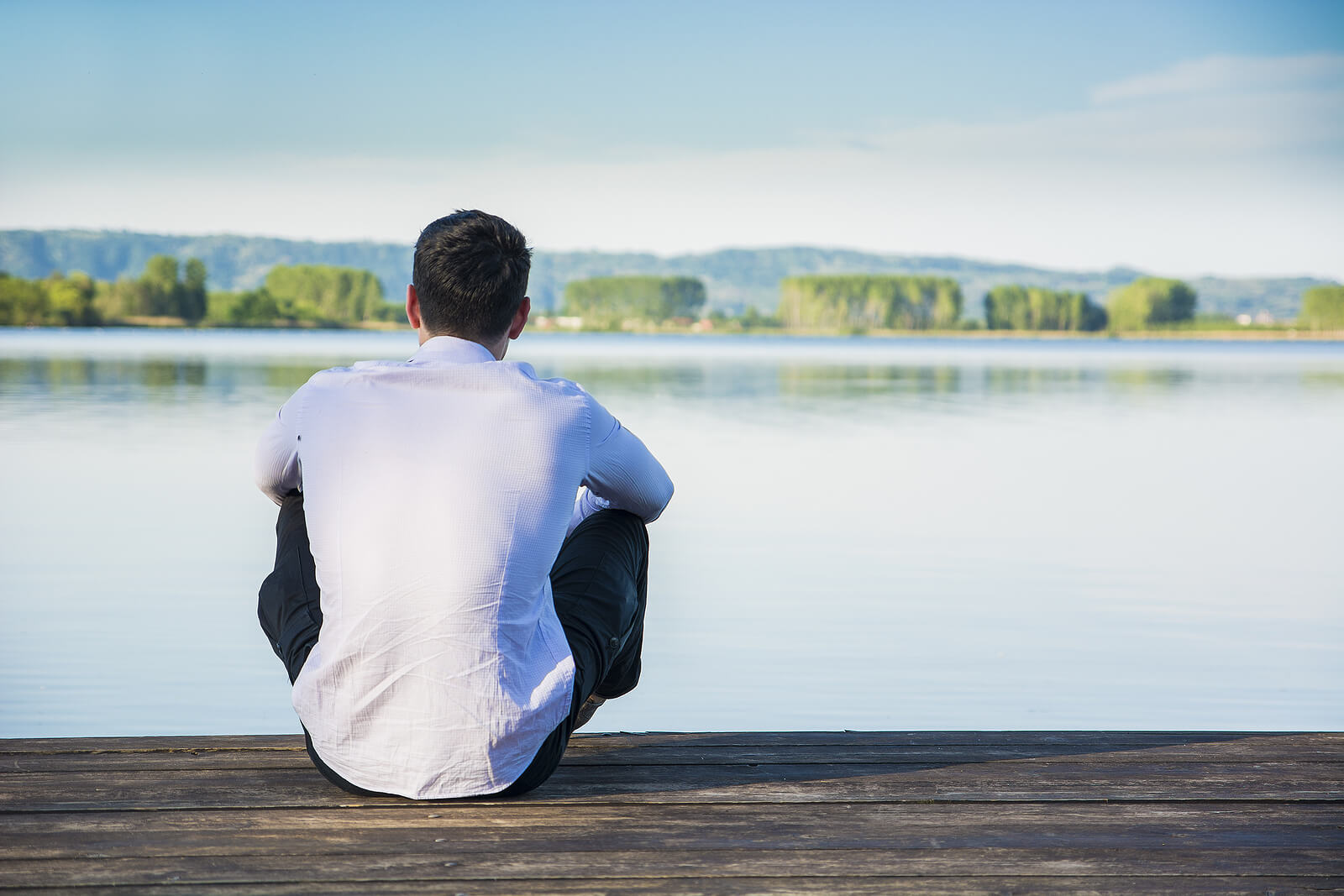 man on dock | emotional strength in mount pleasant, wi | mens therapy | men and mental health | Racine | Burlington | Lake Geneva A man sitting alone on a wooden dock, looking out over a calm lake with trees and hills in the distance. With the support of a men's therapist in Mount Pleasant, WI find emotional balance and find your confidence.