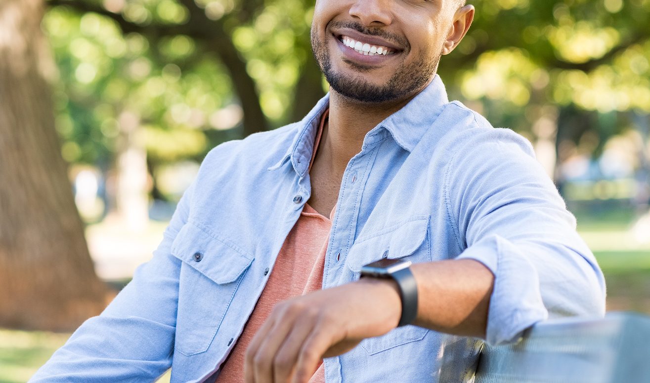 Happy young African man relaxing sitting on bench at a park on a summer day. Overcome your unique mental health struggles and heal in healthy ways with the help of a men's therapist in Kenosha, WI.