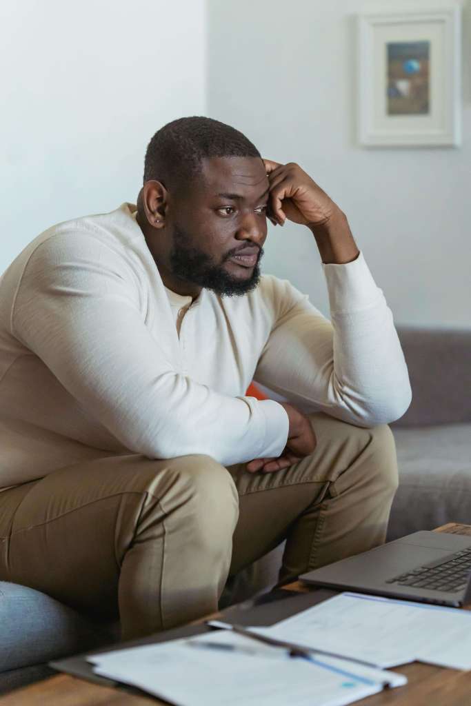 A man sits on a couch leaning forward with his elbow on his knee, looking thoughtfully toward a laptop on a table. Men’s therapy in Milwaukee, WI supports men coping with trauma-related overwhelm, racing thoughts, and internal pressure.