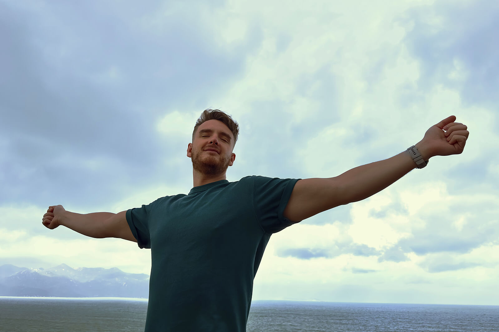 mountains | mens therapy in Milwaukee, WI | mens therapist | mens trauma therapy | Racine | Milwaukee | Madison Man standing with arms outstretched against sky and ocean backdrop. Healing from relational trauma is possible—men's therapy in Milwaukee, WI can help you rebuild trust and reclaim your vitality.
