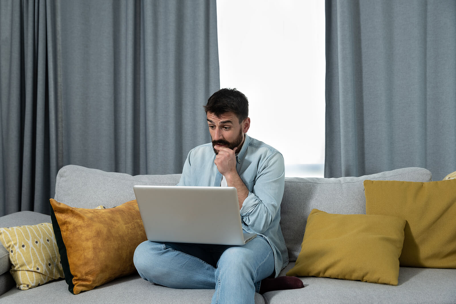 laptop | mens trauma therapy in Milwaukee, WI | mens therapist | high performance men | Racine | Milwaukee | Madison Man sitting on couch with laptop looking thoughtful with hand on chin. When self-doubt and relationship struggles hold you back, men's therapy in Milwaukee, WI provides the support you need to heal.