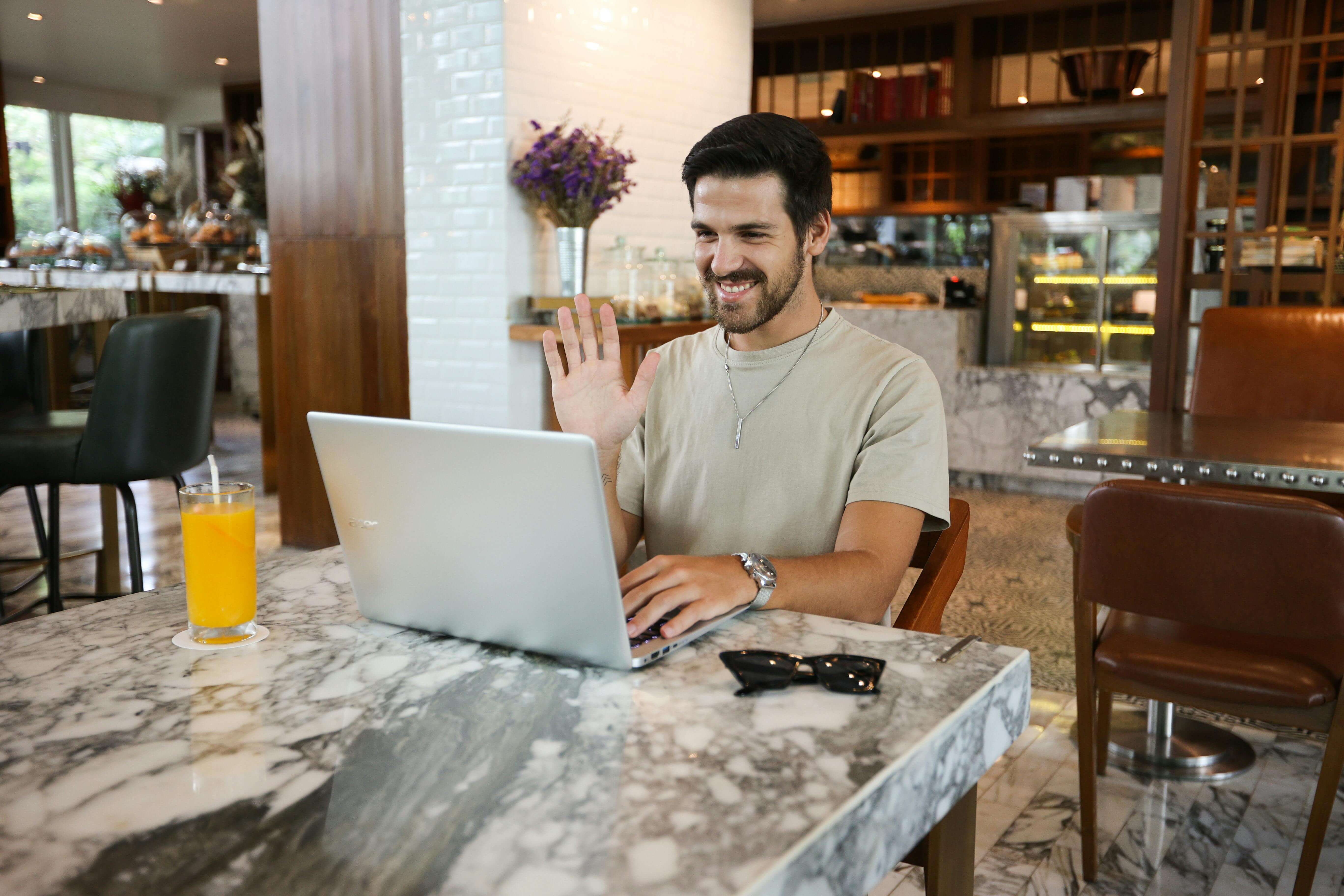 Smiling man waving at laptop screen while sitting at cafe table with orange juice. Men's therapy in Milwaukee, WI helps you achieve your goals without the constant weight of anxiety and sleepless nights.