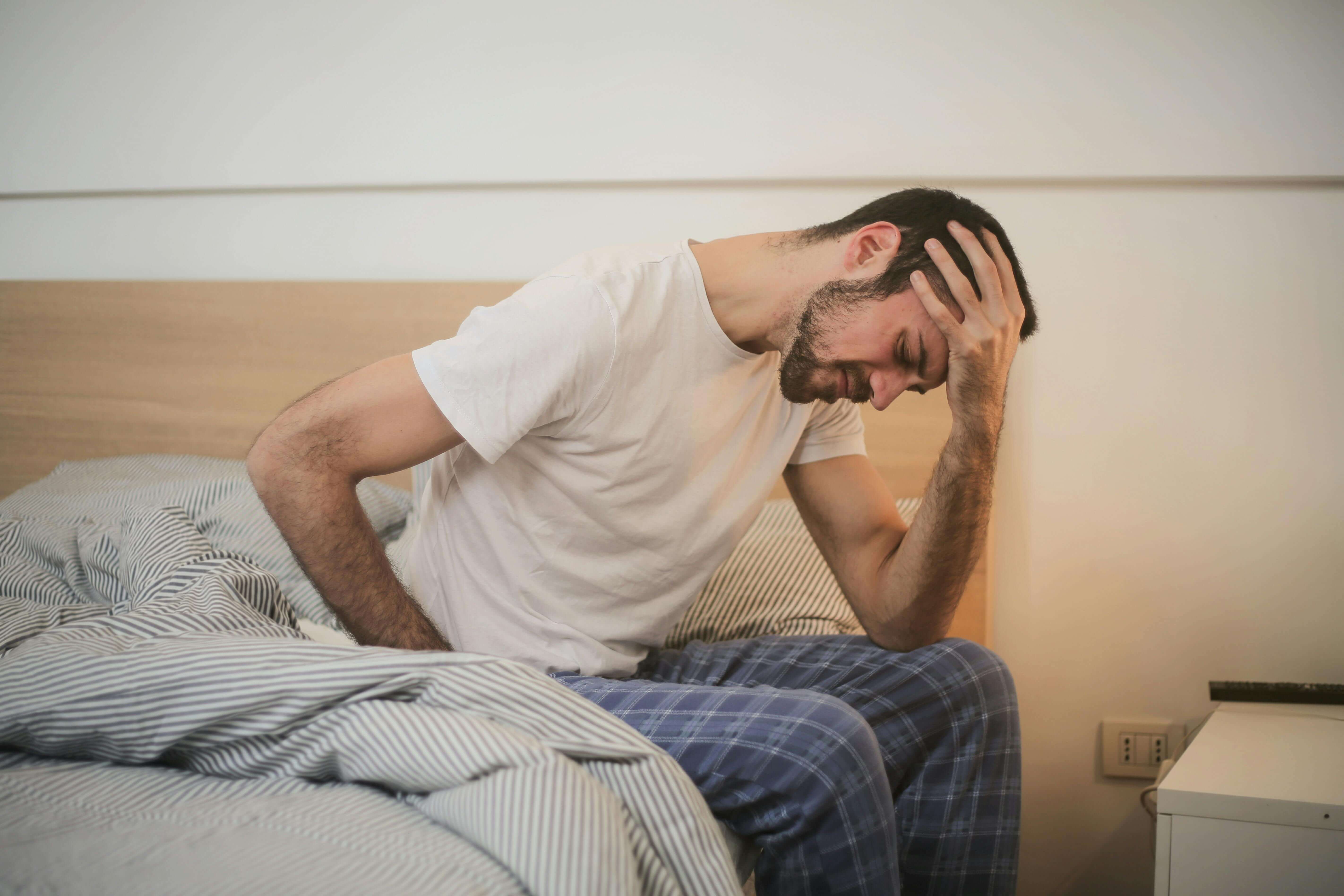 Man in white t-shirt sitting on bed with hand on head looking distressed. Stop going through the motions and start feeling connected to your life with men's therapy in Milwaukee, WI for trauma healing.