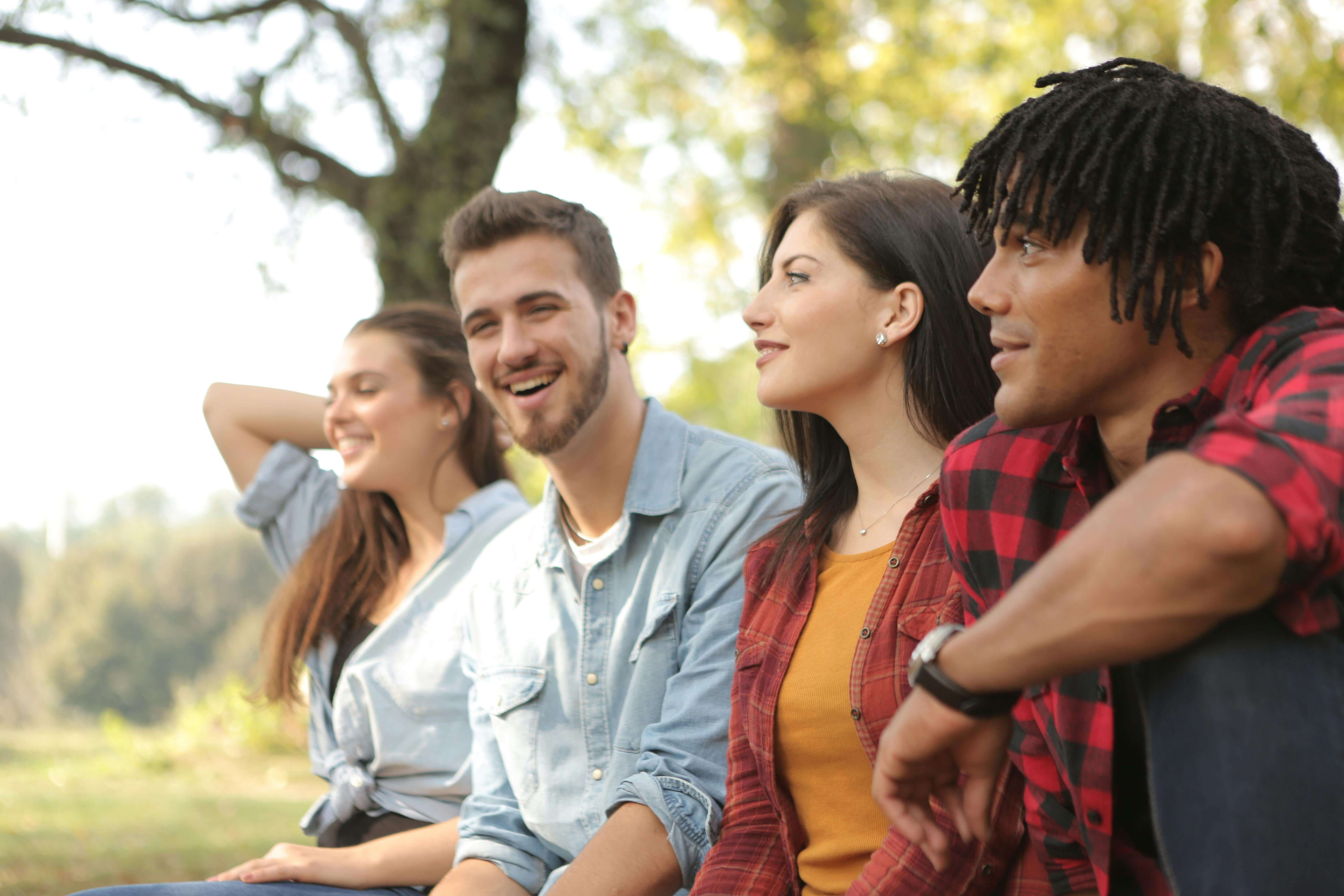 Group of four young adults sitting together outdoors smiling and looking into distance. Teach your nervous system that success and connection are safe to keep with men's trauma therapy in Milwaukee, WI.
