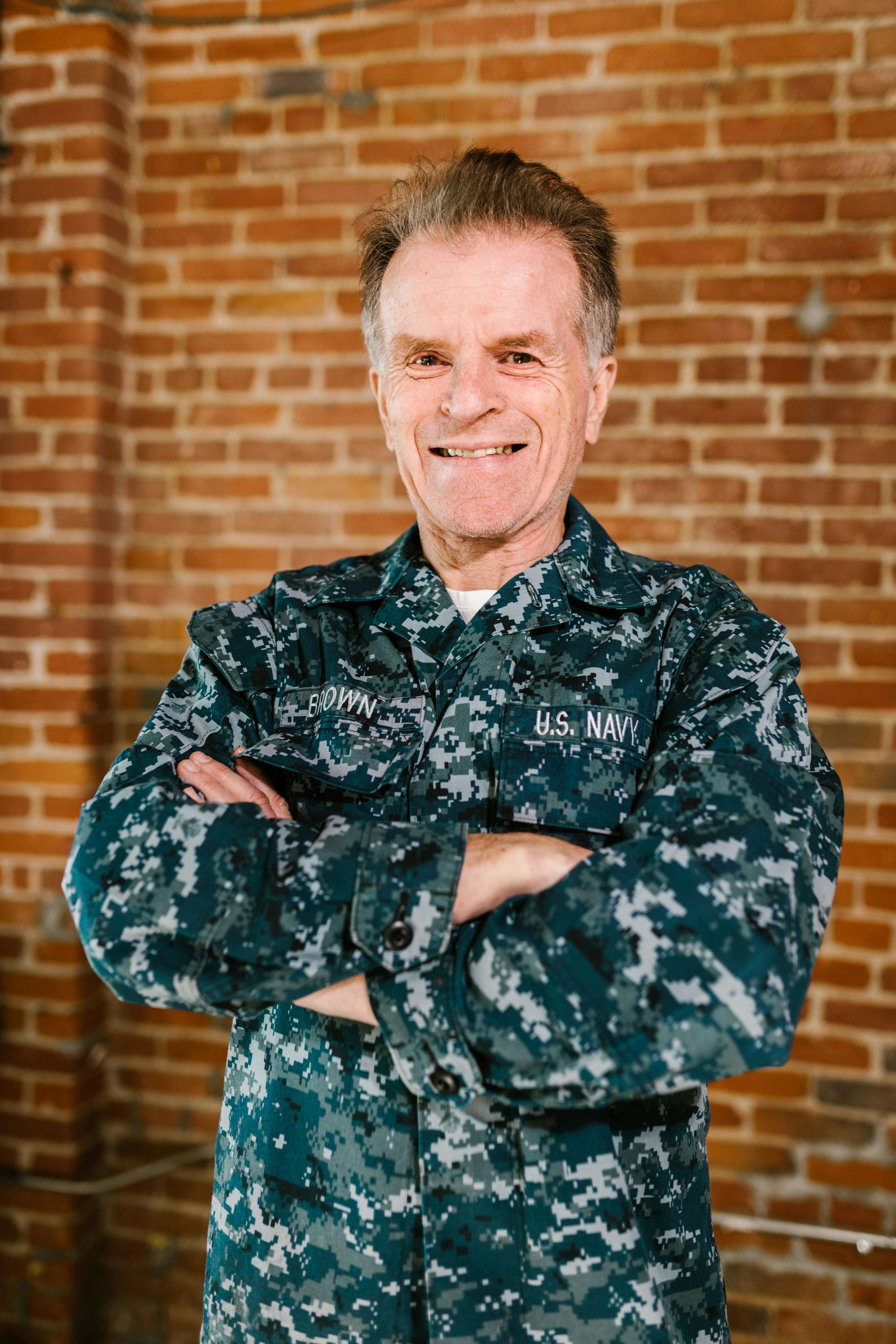 Smiling U.S. Navy service member in digital camouflage uniform with arms crossed against brick wall. Process trauma and regulate your nervous system with specialized men's therapy in Milwaukee, WI.