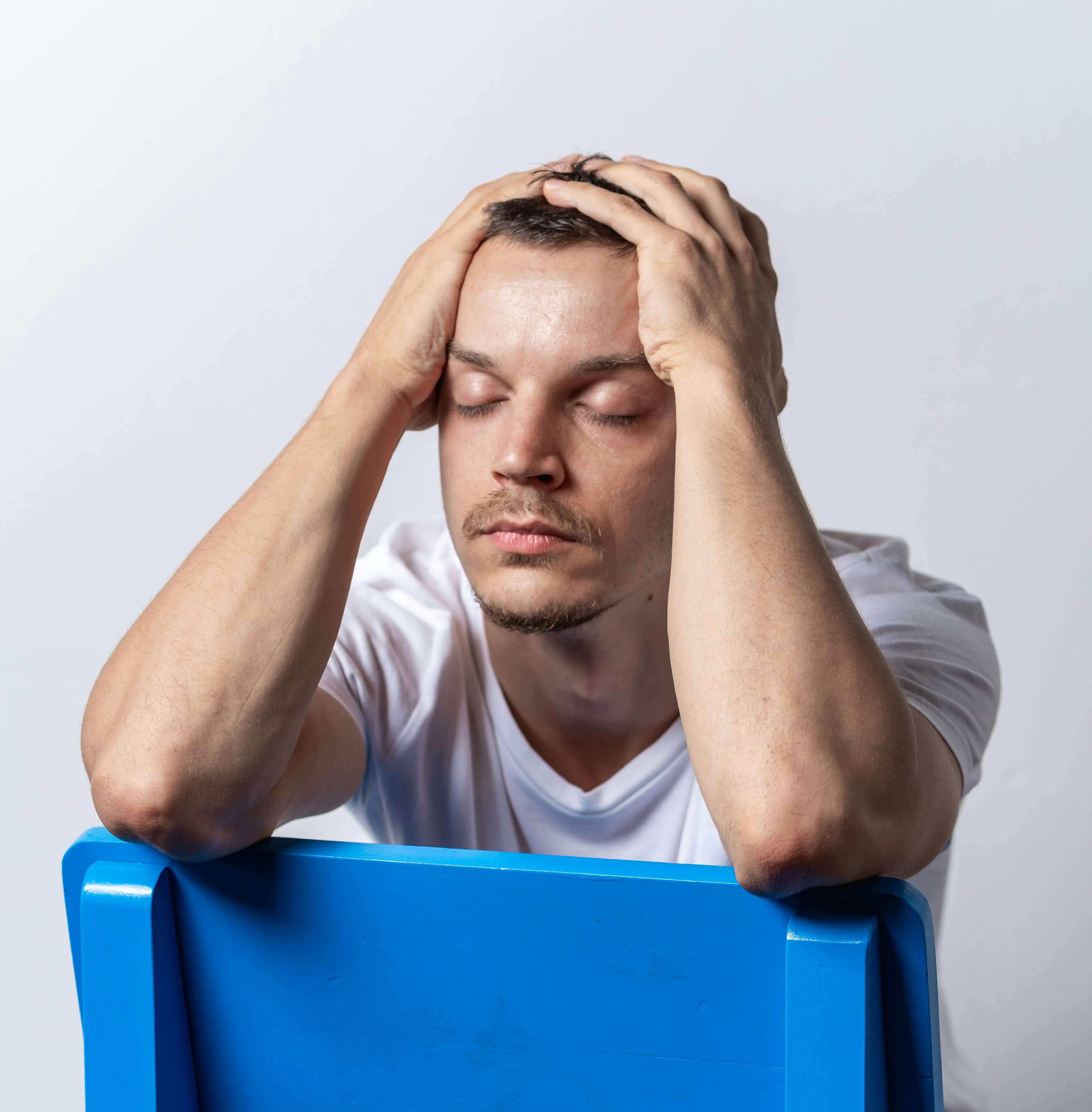 Man in white v-neck shirt with both hands on head sitting in blue chair with eyes closed. Stop living on autopilot and rebuild your mind-body connection with men's therapy in Milwaukee, WI.