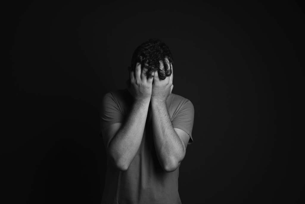 Black and white photo of man in t-shirt with both hands covering face against dark background. Break through emotional numbness and reconnect with your feelings through men's therapy in Milwaukee, WI.
