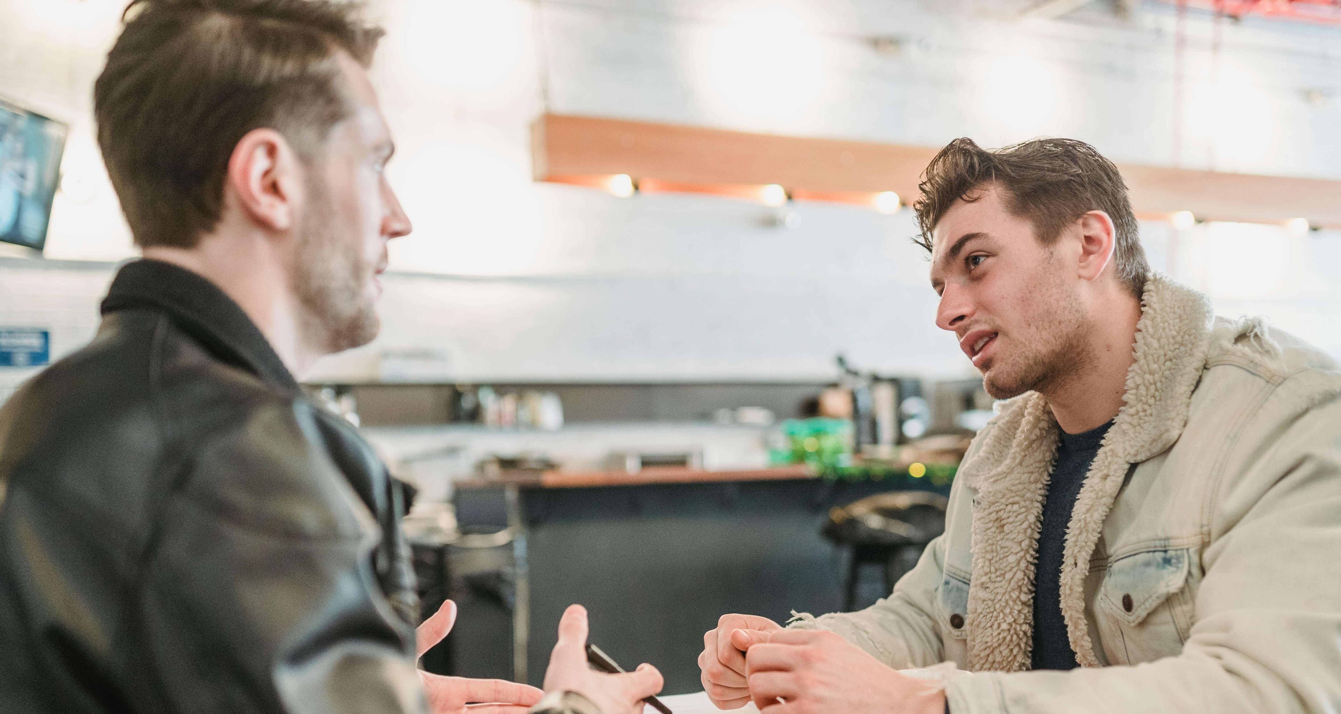 Two men having conversation at table in bright modern space with one gesturing while talking. Process unresolved trauma and learn to feel emotions again through men's therapy in Milwaukee, WI.