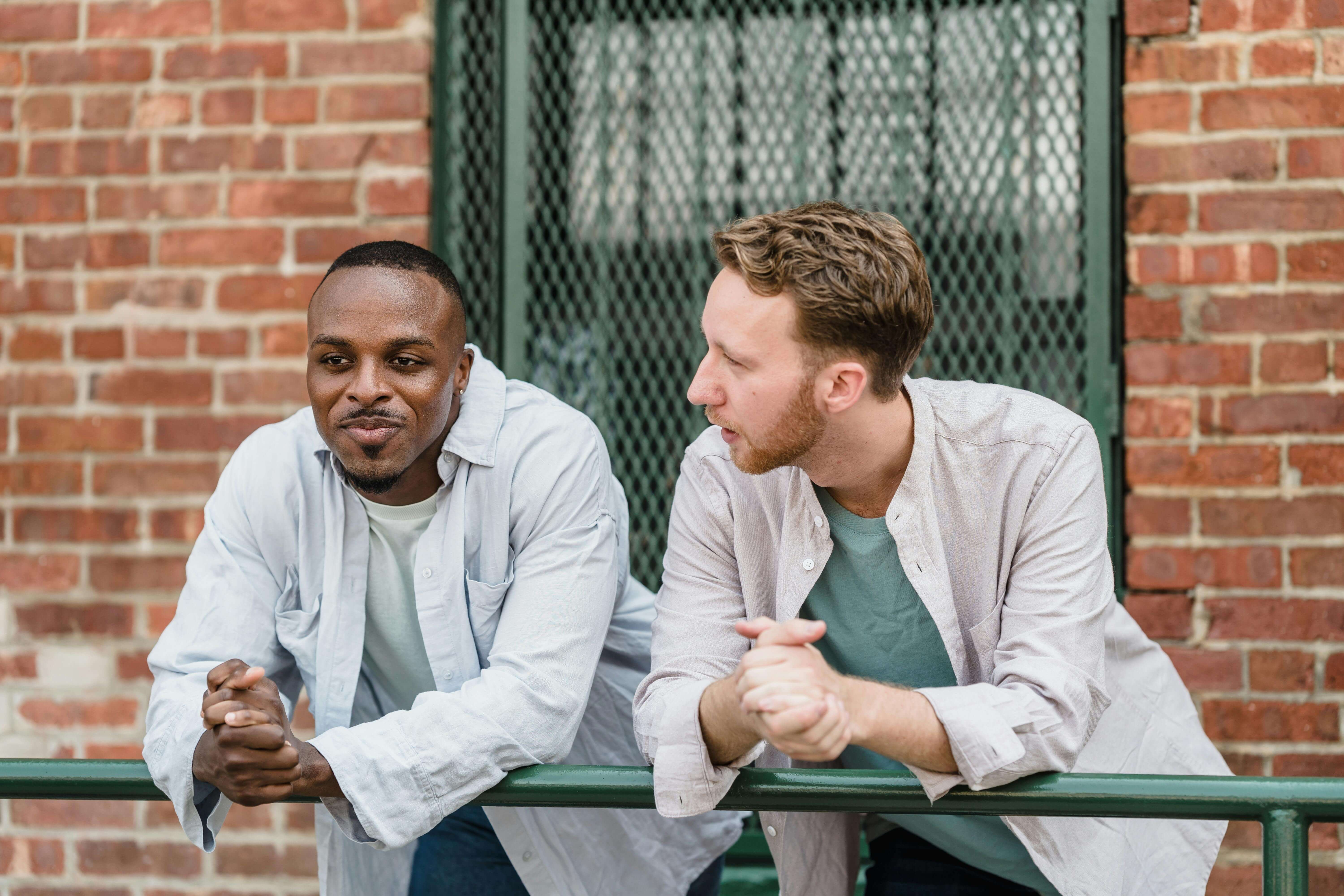 Two men in button-up shirts sitting on green bench having conversation outdoors near brick building. Overcome emotional disconnection and feel alive again with somatic approaches in men's therapy in Milwaukee, WI.