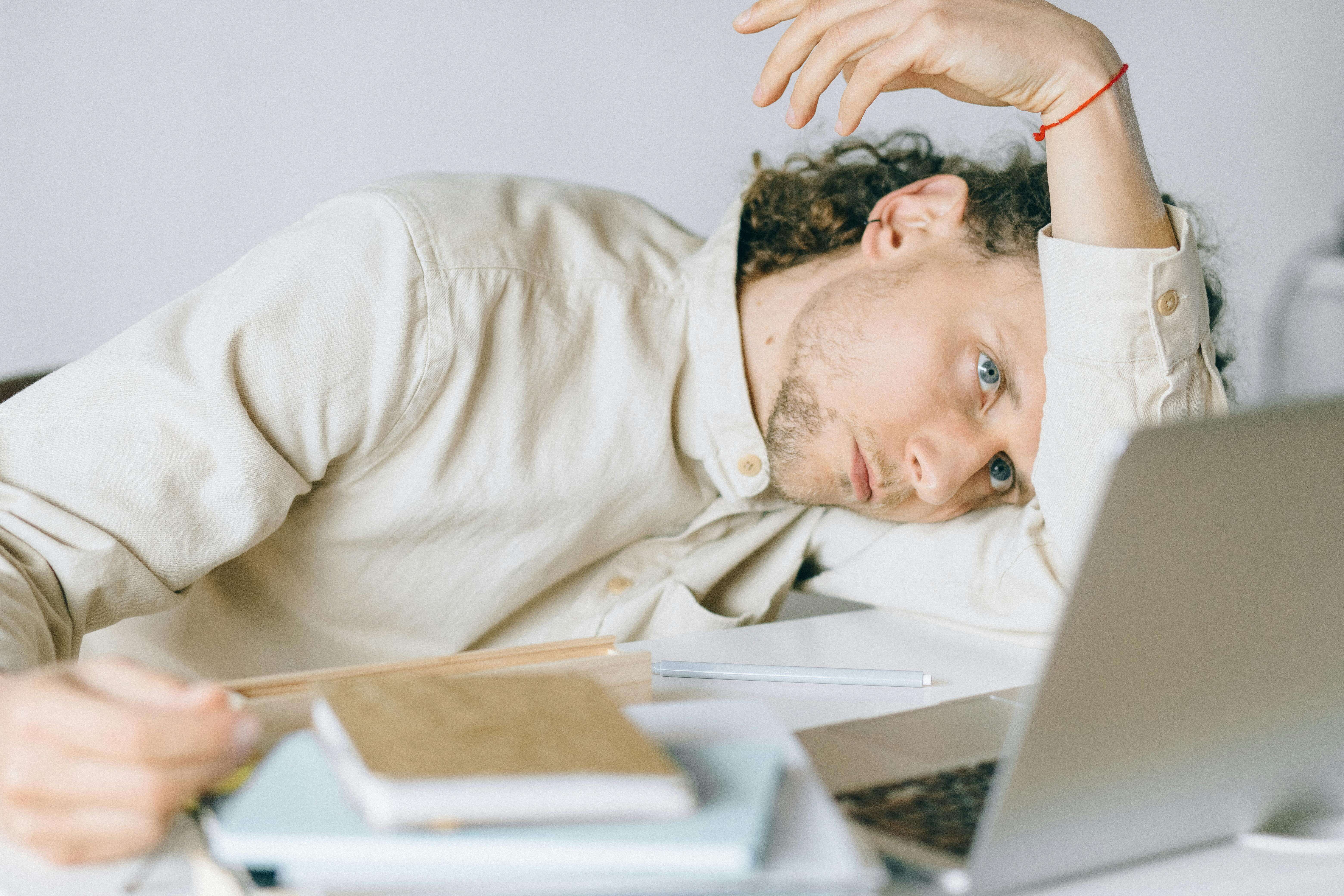 Man in cream colored shirt lying on desk looking tired with laptop and papers nearby. Stop carrying the weight of business collapse and unresolved trauma alone through men's therapy in Milwaukee, WI.