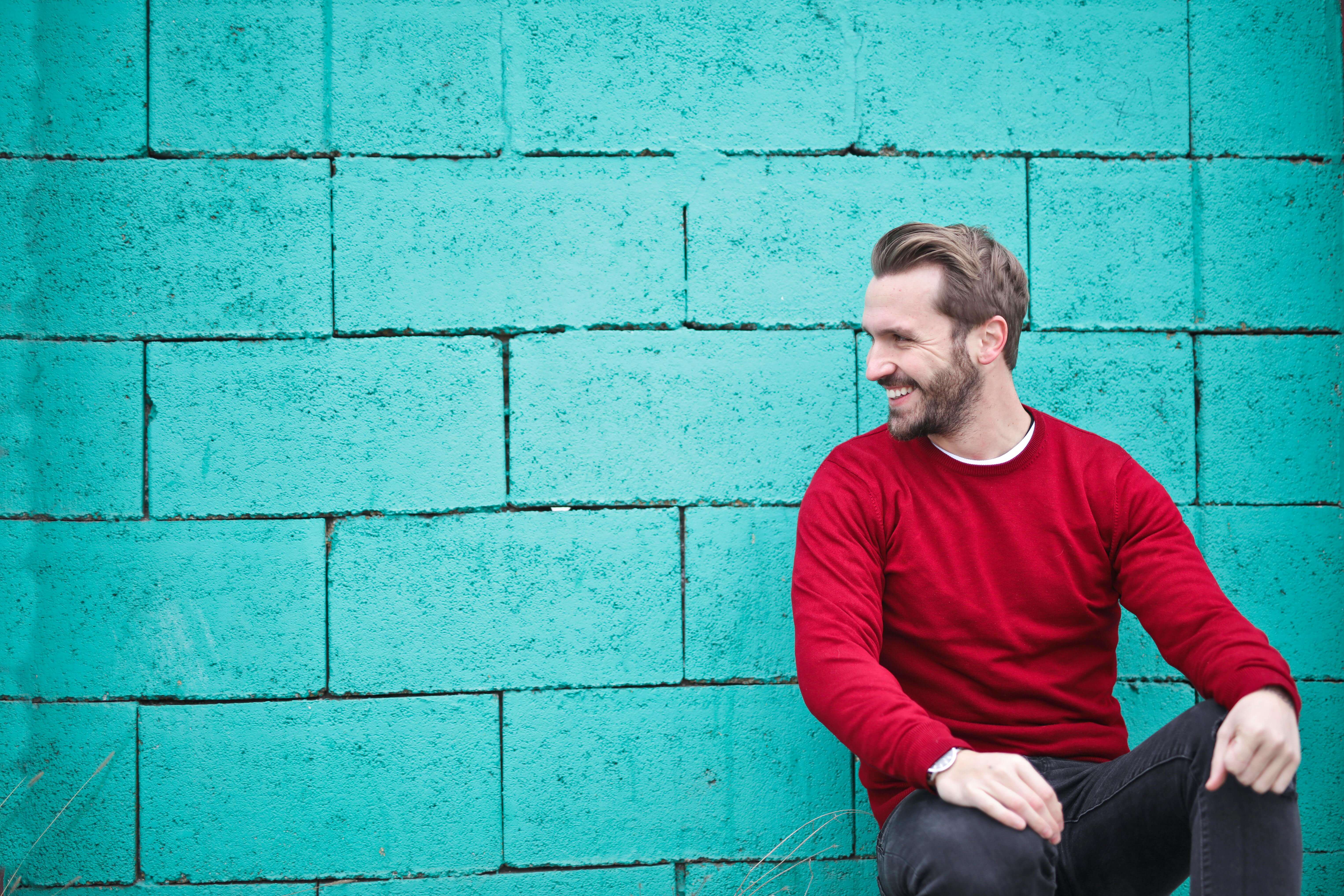 brick wall | mens therapy in Milwaukee, WI | performance anxiety | mens anxiety | Mount Pleasant | Burlington | Lake Geneva Man in red sweater smiling while sitting against bright turquoise brick wall. Learn to perform without overreacting or shutting down and reclaim your confidence with men's therapy in Milwaukee, WI.