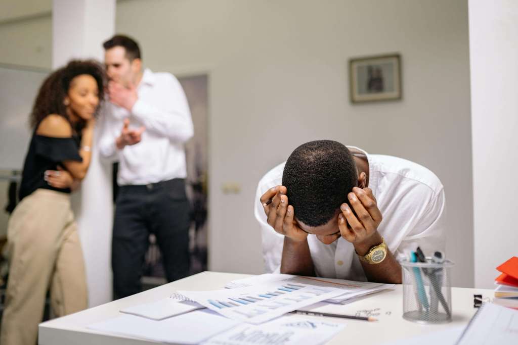 Man in white dress shirt with head in hands at desk with charts while colleagues argue in background. Stop freezing before critical moments and rebuild your performance confidence with men's therapy in Milwaukee, WI.