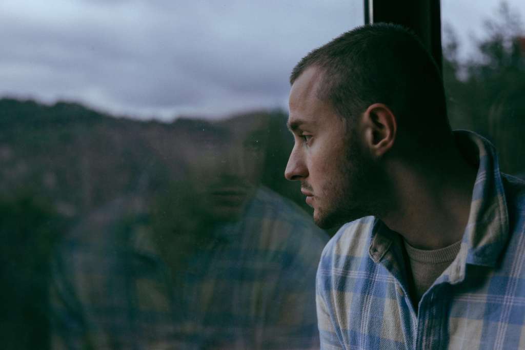 A man in a blue plaid shirt looking out a rain-streaked window, his reflection visible in the glass against a cloudy, wooded background. You don't have to keep staring down the silence alone — men's therapy in Milwaukee, WI offers a path toward healing and calm.