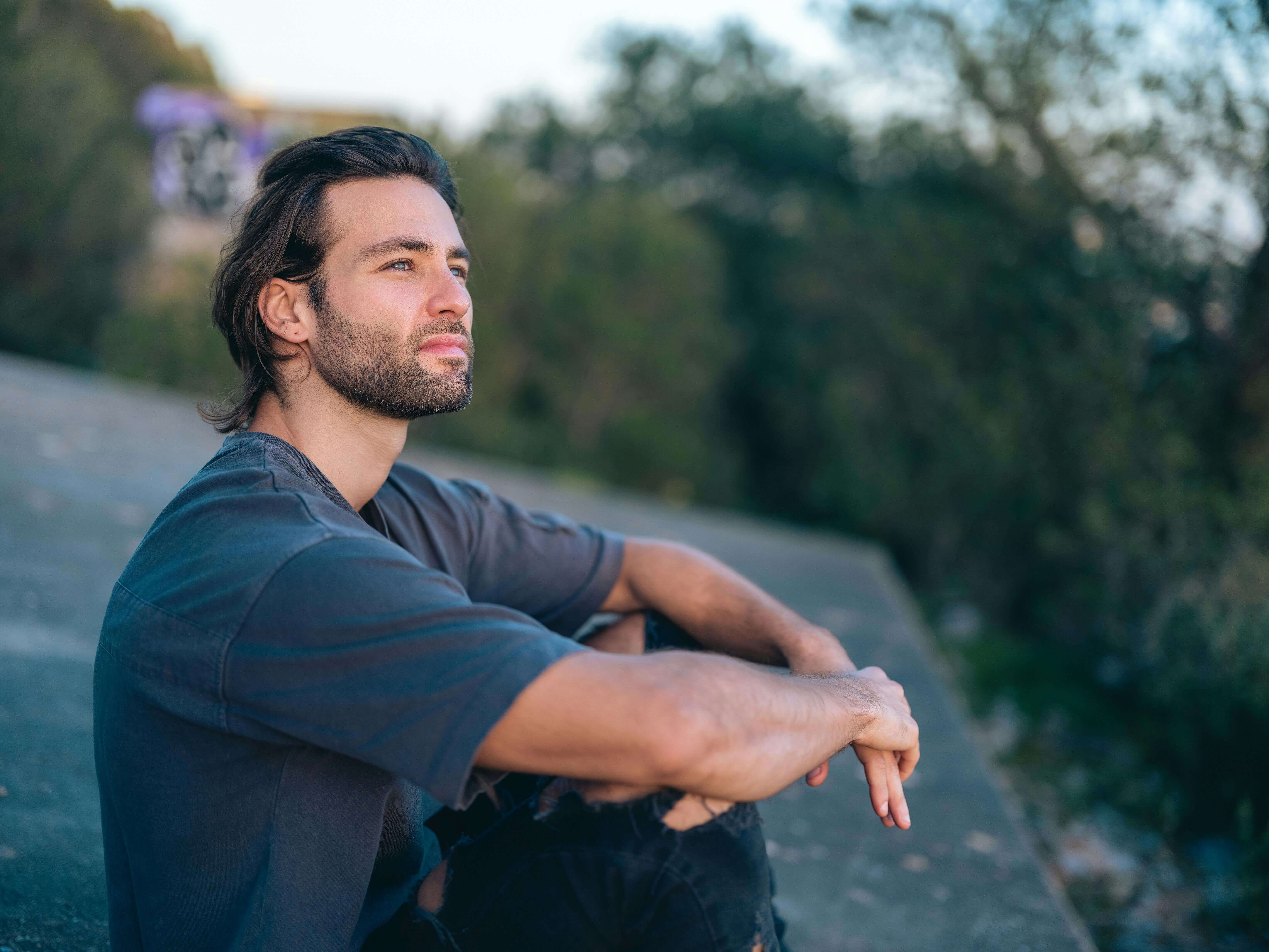 young man sitting outside | mens therapy in Milwaukee, WI | men and mental health | mens anxiety, men and depression | Pewaukee | Greenfield | Greendale A young man with dark hair sitting on a concrete surface outdoors, arms resting on his knees, gazing thoughtfully into the distance. Through men's therapy in Milwaukee, WI, small-town men can finally release the emotional burden they've been silently holding.