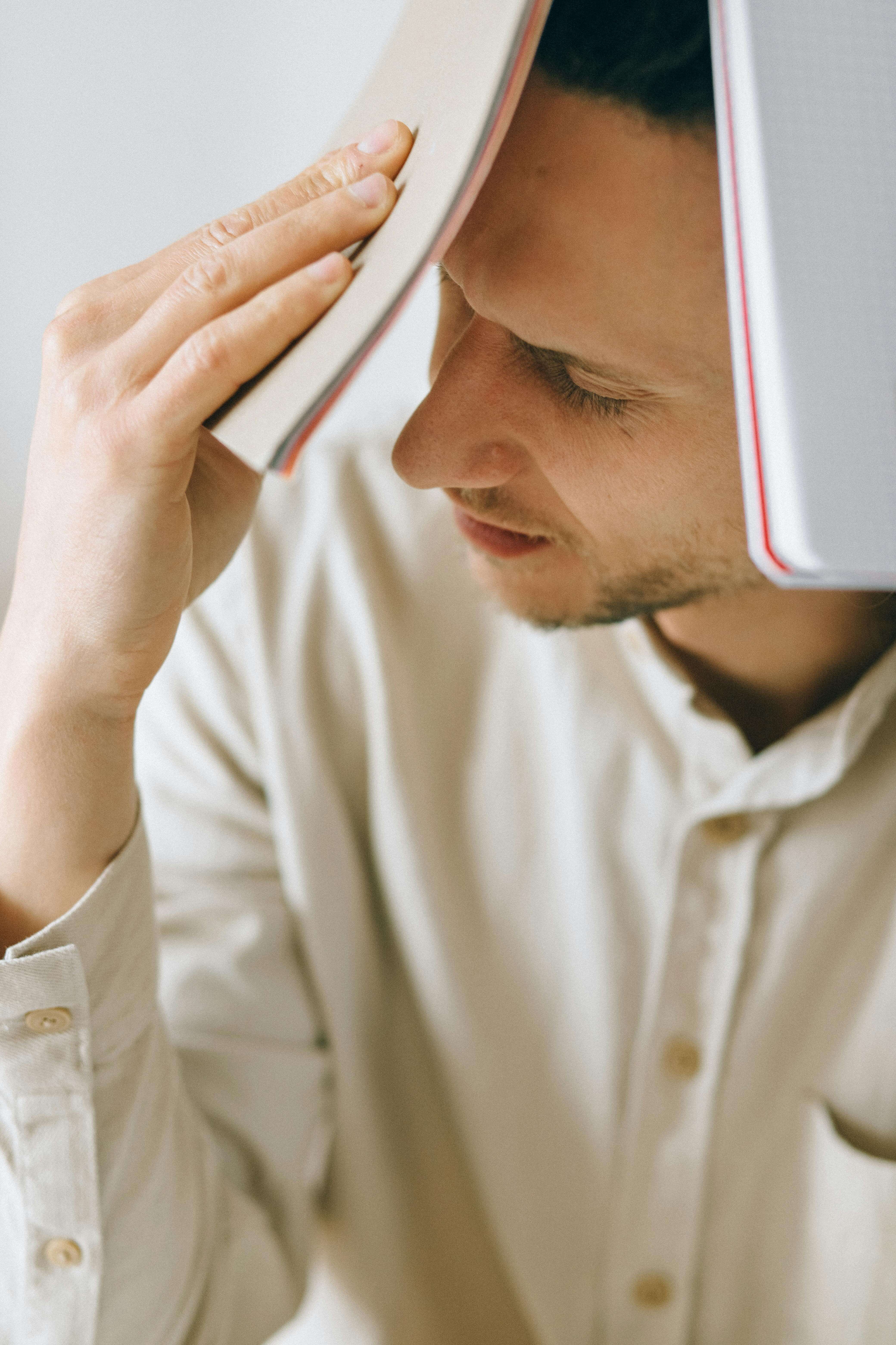 Man in cream button-up shirt holding book against forehead with eyes closed looking down. Address physical symptoms like headaches, chest pain, and fatigue caused by anxiety and depression with men's therapy in Milwaukee, WI.