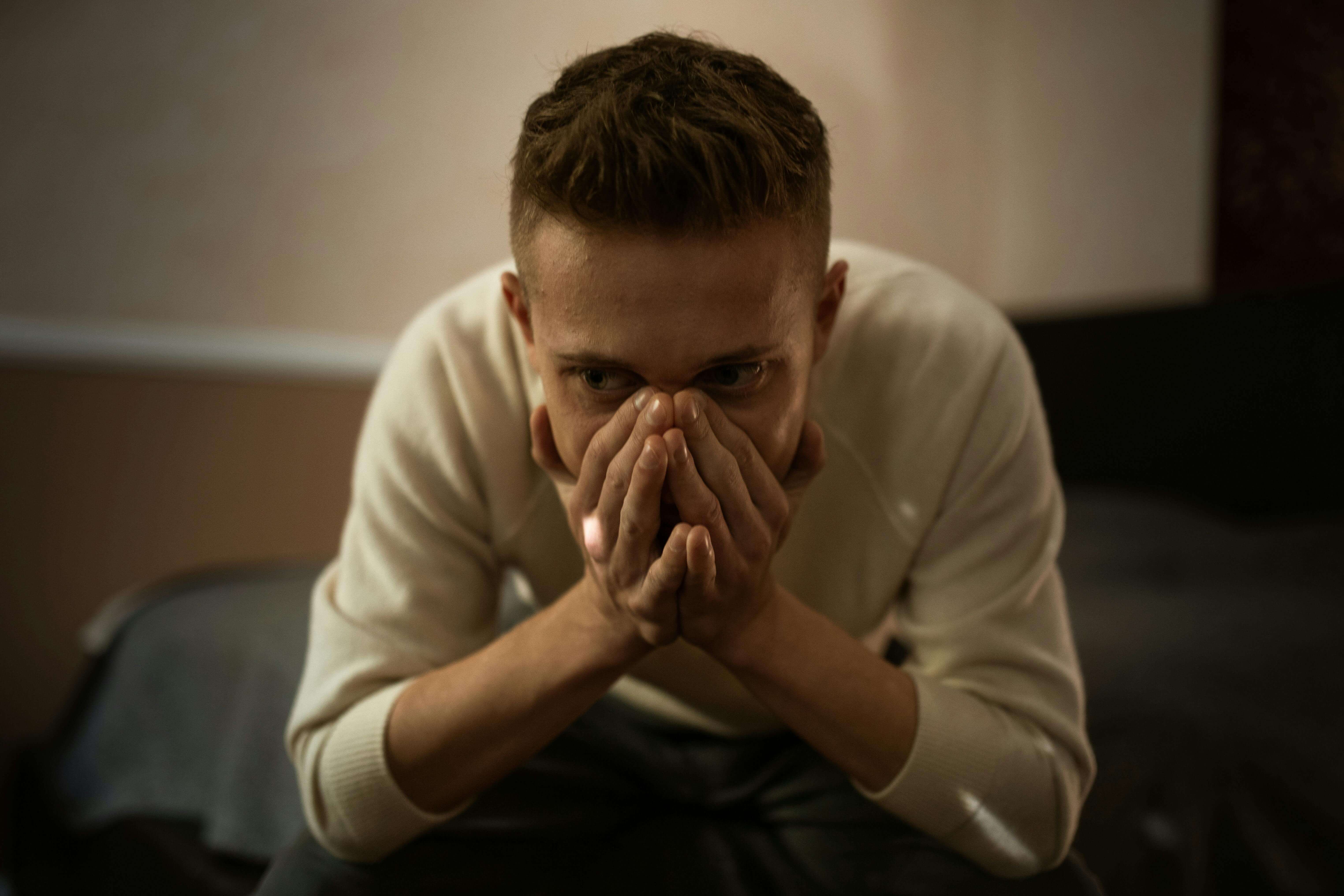 Man in beige sweater sitting with clasped hands covering lower face looking worried in dim lighting. Stop getting told "nothing is wrong" and find relief from the physical toll of anxiety and depression through men's therapy in Milwaukee, WI.