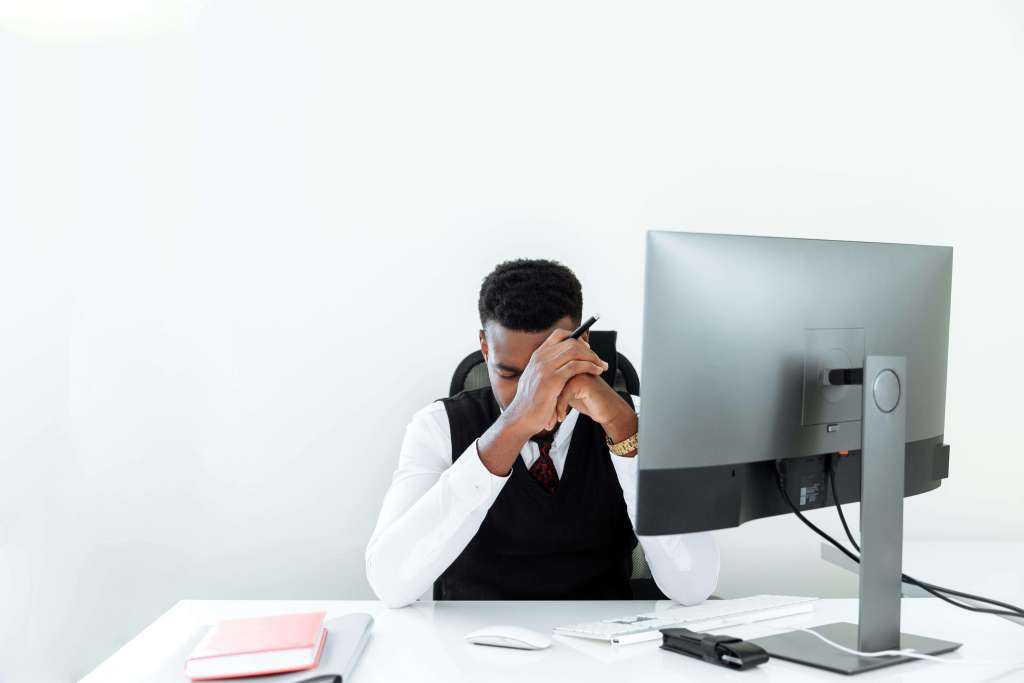 Man in black vest and white shirt sitting at desk with head down holding forehead looking stressed at computer. Stop carrying the weight of financial responsibility and decision fatigue alone with men's therapy in Milwaukee, WI designed for entrepreneurs.