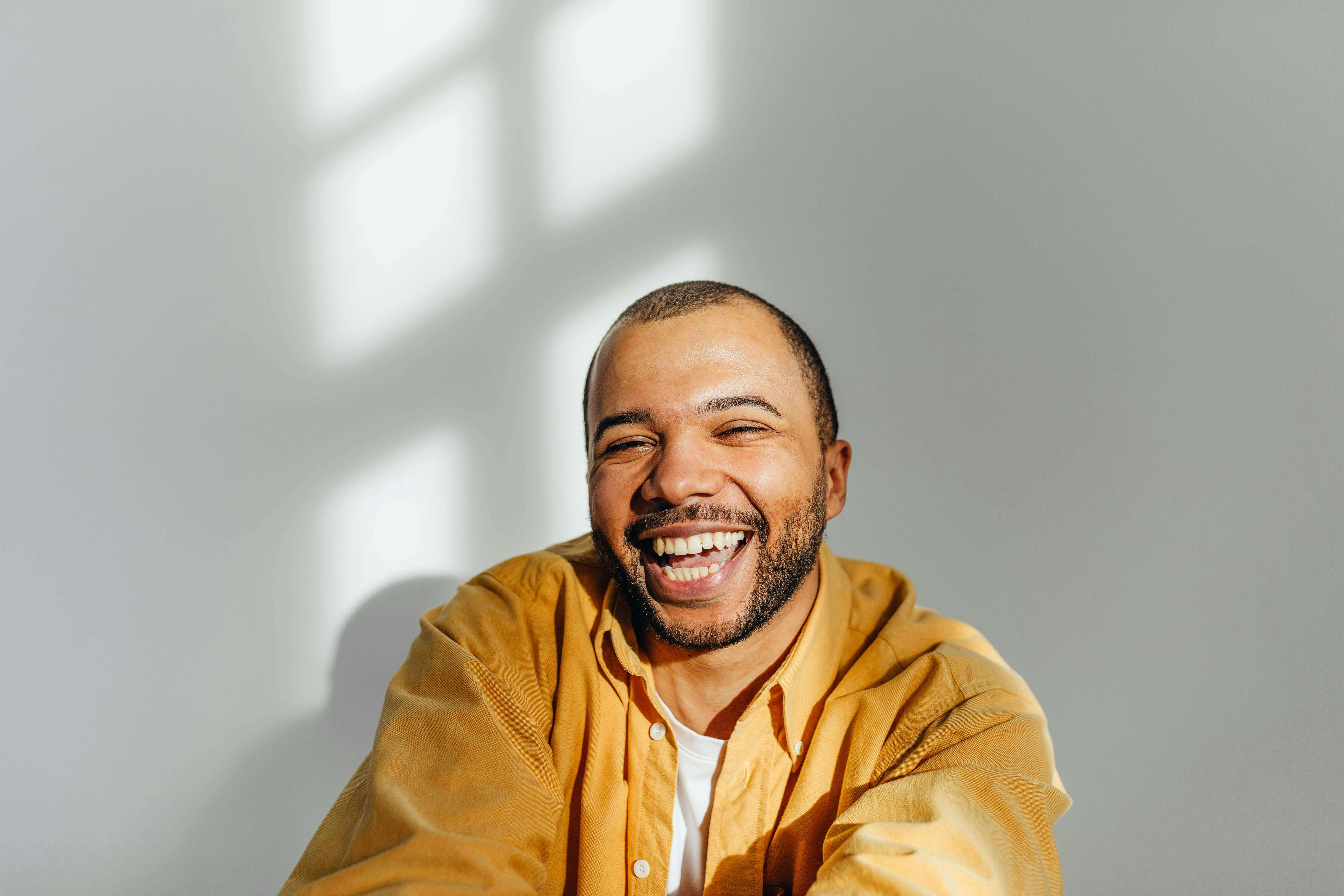 Man in mustard yellow hoodie smiling broadly against white wall with window shadow. Find lasting relief from chronic fatigue, muscle tension, and sleep problems by addressing the root cause with men's therapy in Milwaukee, WI.