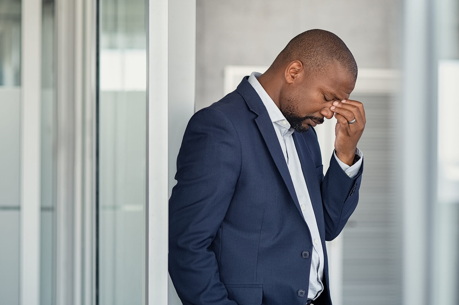 man in suit | men's therapy in Milwaukee, WI | men's anxiety | men and mental health | Elm Grove | Wauwatosa | Brookfield Man in a navy suit leaning against a wall in an office corridor with his hand pressed to his face and his head down. When anxiety follows you into the boardroom and beyond, men's therapy in Milwaukee, WI helps you finally break the cycle.