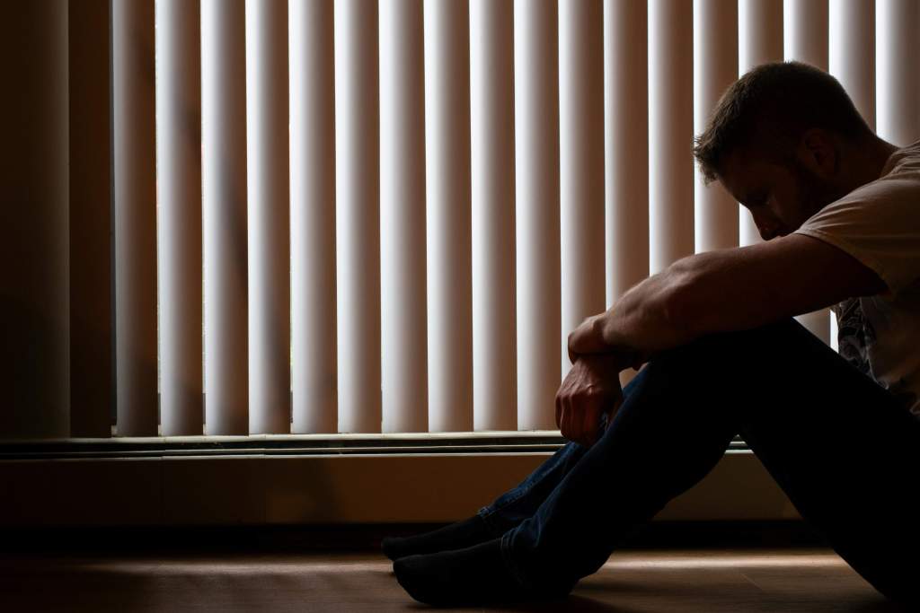 Man sitting on the floor with his back against window blinds and his head down in the shadows. You don't have to sit alone with the weight of loss — men's therapy for grief in Milwaukee, WI can help you find your way through.
