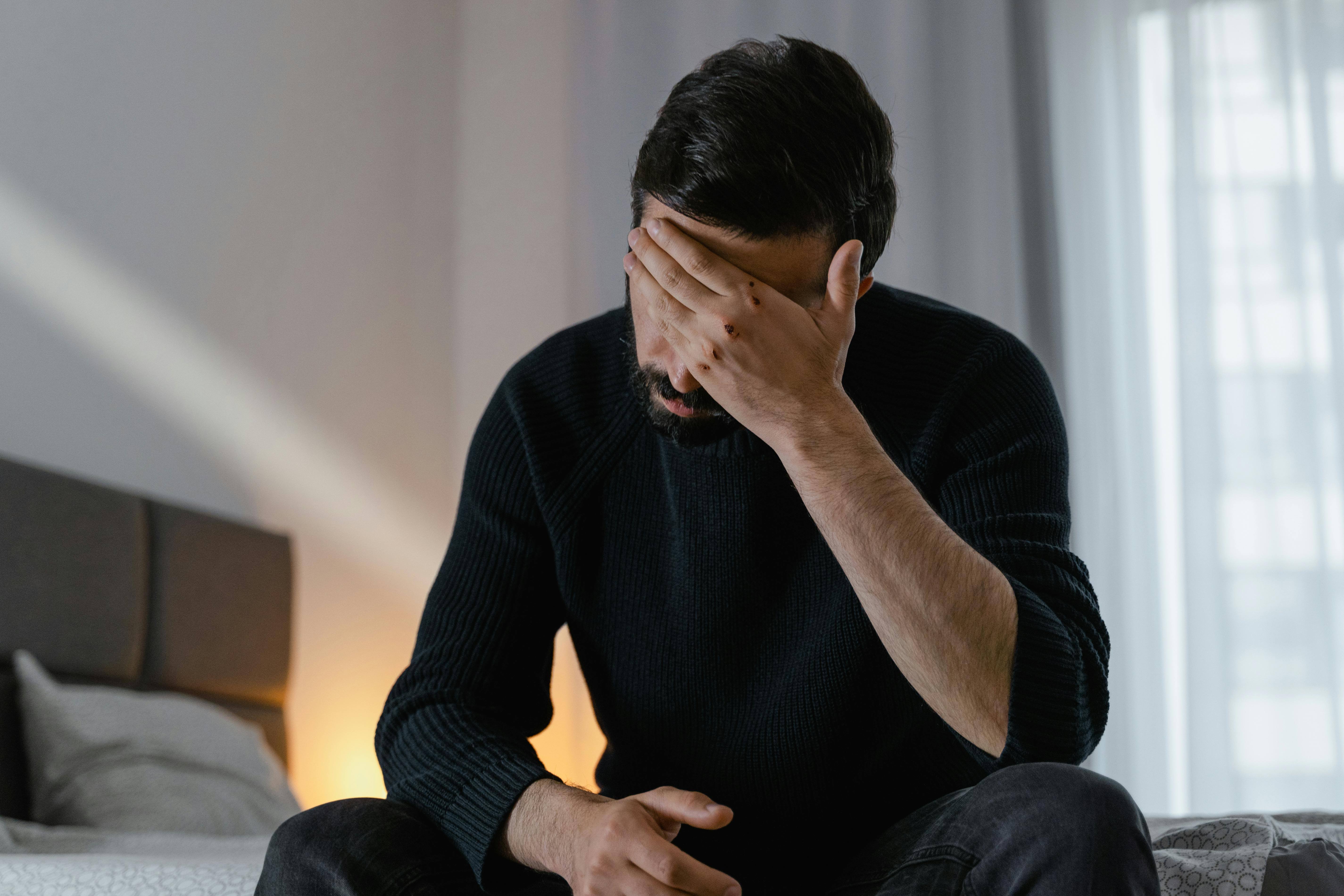 Bearded man in a black sweater sitting on a bed with one hand covering his face in a dimly lit room. When trauma starts affecting every part of your life, men's trauma therapy in Milwaukee, WI can help you reclaim your sense of self.