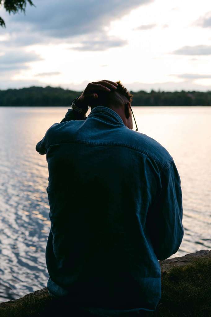 Man in a denim jacket sitting on a rock with his hand on his head facing a lake at dusk. You don't have to keep carrying it alone — men's trauma therapy in Milwaukee, WI gives you the tools to process what your body has been holding.