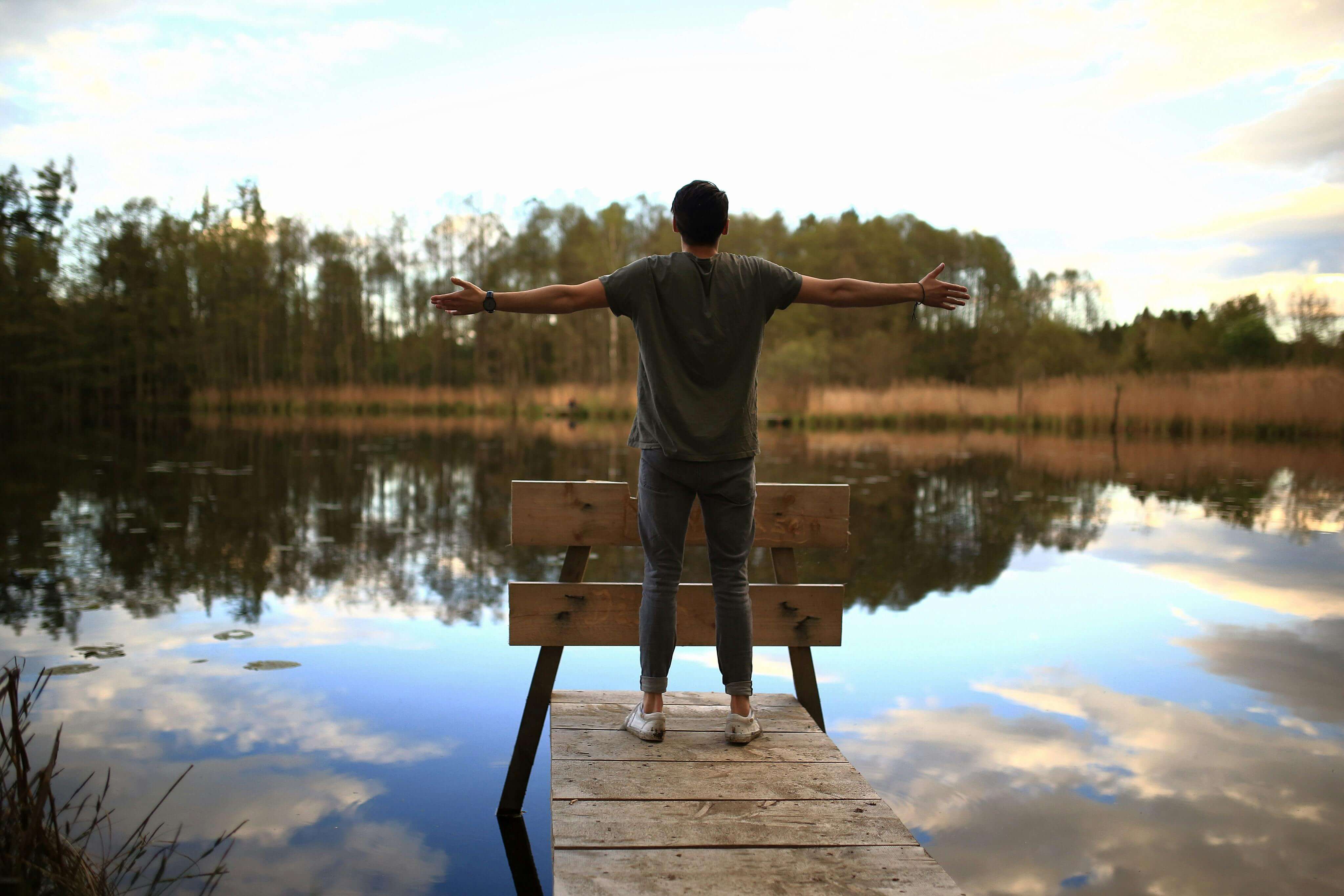 man standing on dock | men's therapy in Milwaukee, WI | men with anxiety and depression | men and mental health | Oak Creek | River Hills Man standing on a wooden dock with his arms stretched wide open facing a calm lake surrounded by trees and clouds. Calm is possible with the support of men's therapy in Milwaukee, WI. It can help you regulate your nervous system and reconnect with the peace anxiety has taken from you.