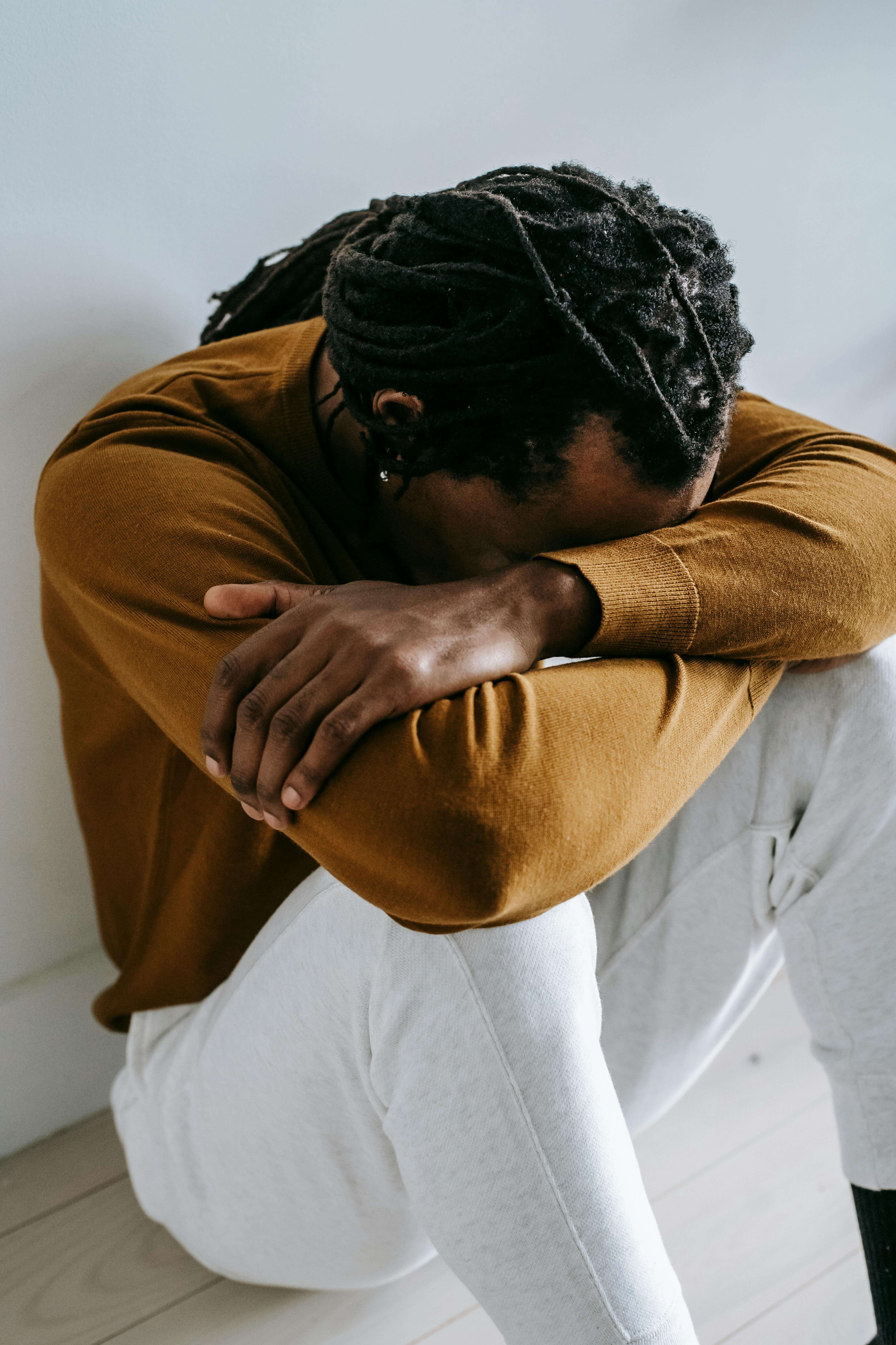 man sitting on floor | trauma therapy in Milwaukee, WI | men and mental health | trauma therapy for men | Elm Grove | Wauwatosa | Brookfield Man with locs wearing an orange sweater sitting on the floor with his head buried in his arms against a white wall. You don't have to keep carrying the weight of the past alone. Trauma therapy in Milwaukee, WI helps men finally release what the body has been holding.