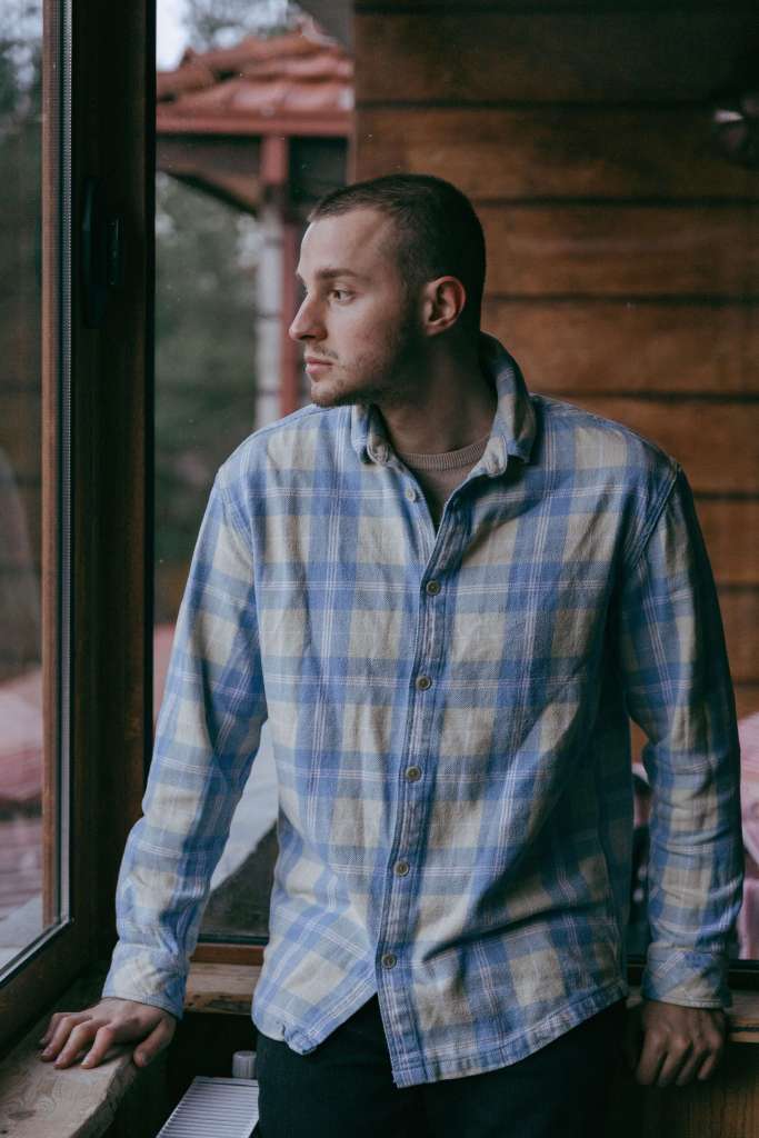 Young white man with short hair wearing a blue plaid flannel shirt leaning against a window frame, looking outside with a distant expression. If you've been carrying weight you can't quite name, trauma therapy in Milwaukee, WI can help you finally understand what your nervous system has been holding.