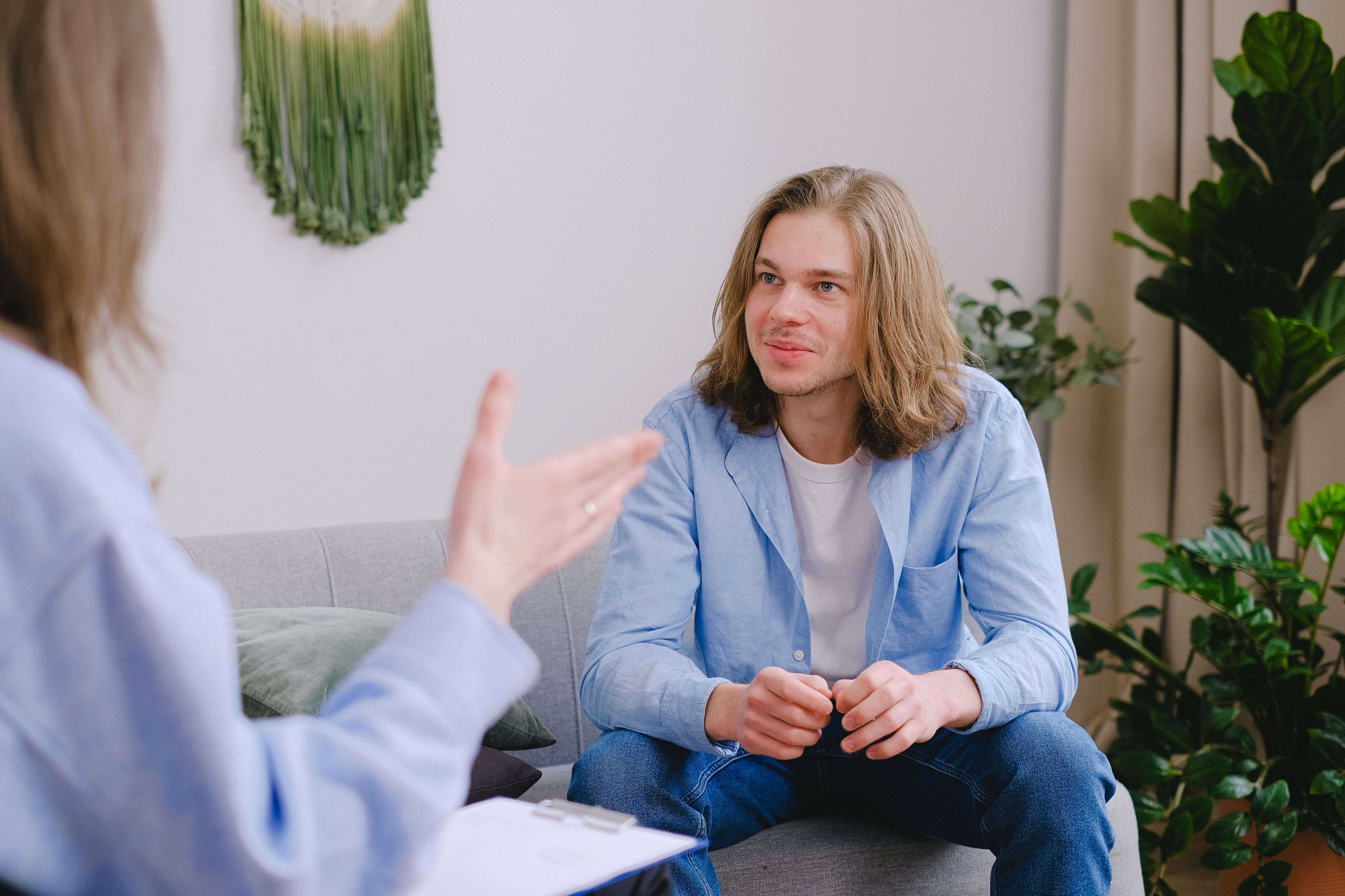 Young man with long blonde hair sitting on a gray couch in a plant-filled therapy office, listening attentively to a therapist gesturing in the foreground. The right therapeutic relationship makes all the difference and trauma therapy in Milwaukee, WI is collaborative, body-forward, and paced to meet you where you are.