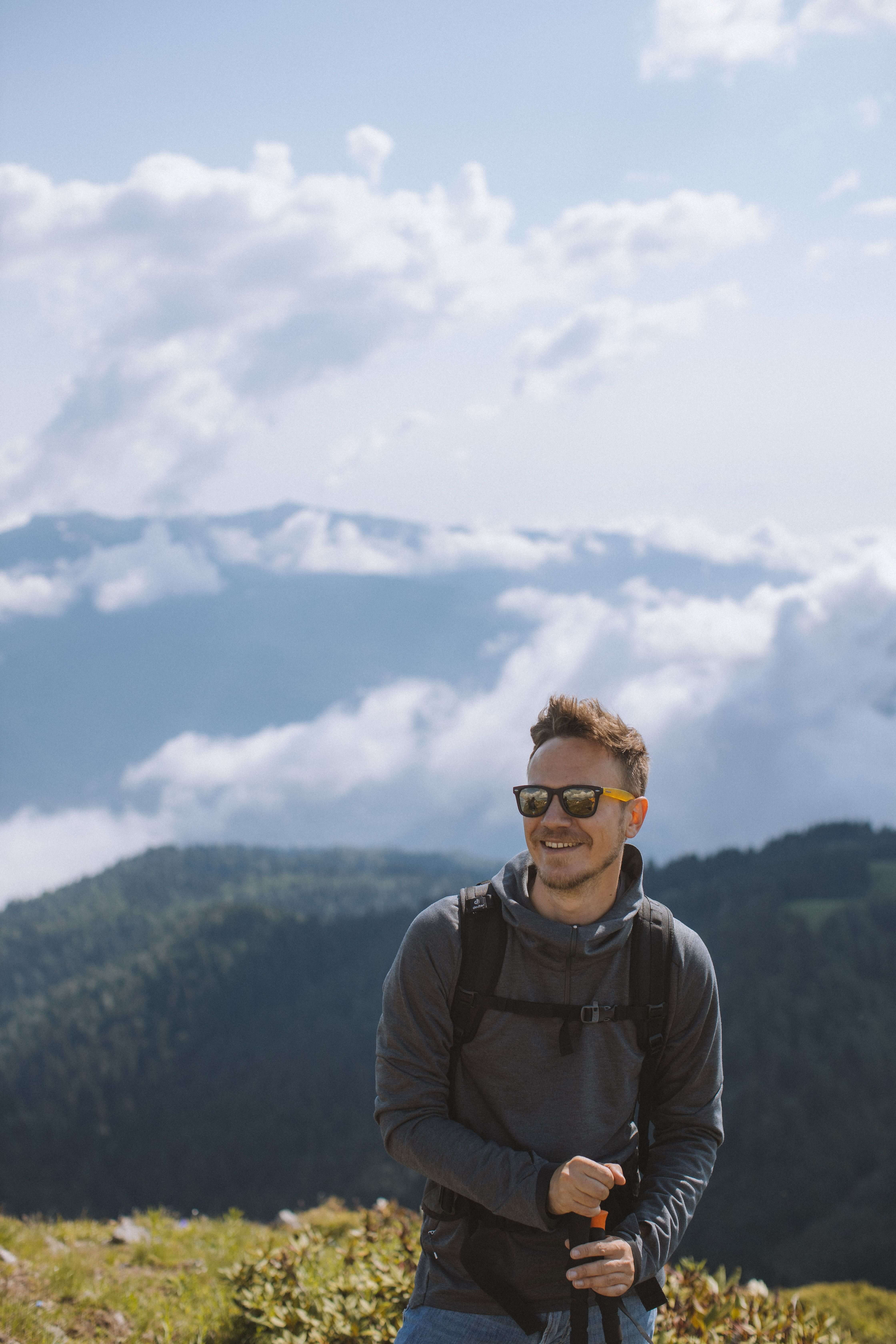 Smiling man wearing sunglasses and a backpack standing outdoors with mountains and clouds behind him. Healing from trauma doesn't mean losing yourself. Trauma therapy in Milwaukee, WI helps men reclaim their confidence and their life.