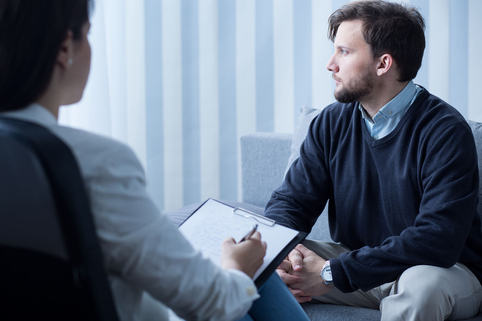 Bearded man in a navy sweater sitting across from a therapist who is taking notes on a clipboard in a softly lit therapy office. Taking the first step toward healing takes courage. Trauma therapy in Milwaukee, WI gives men a safe and confidential space to begin recovering from betrayal trauma.