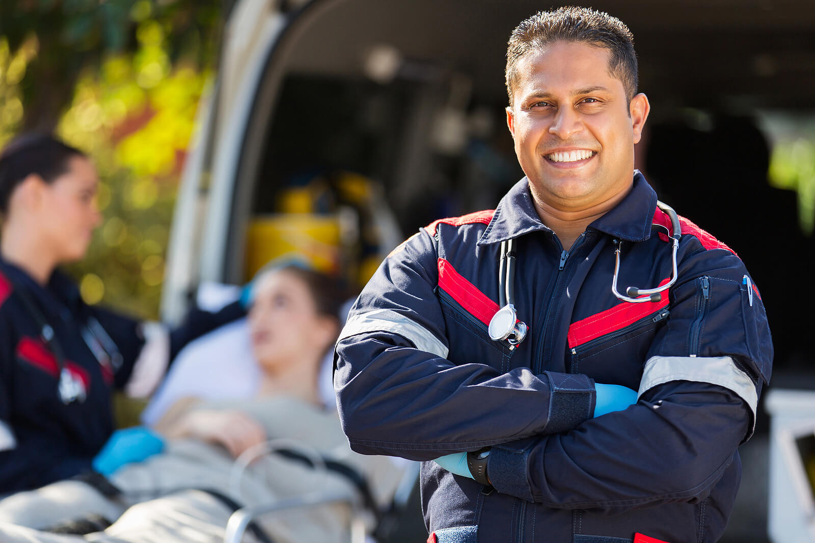 Male paramedic in uniform smiling with arms crossed in front of an ambulance with a patient and colleague in the background. Strength on the job doesn't mean you have to carry it alone. Trauma therapy in Milwaukee, WI helps first responders heal without leaving their identity behind.