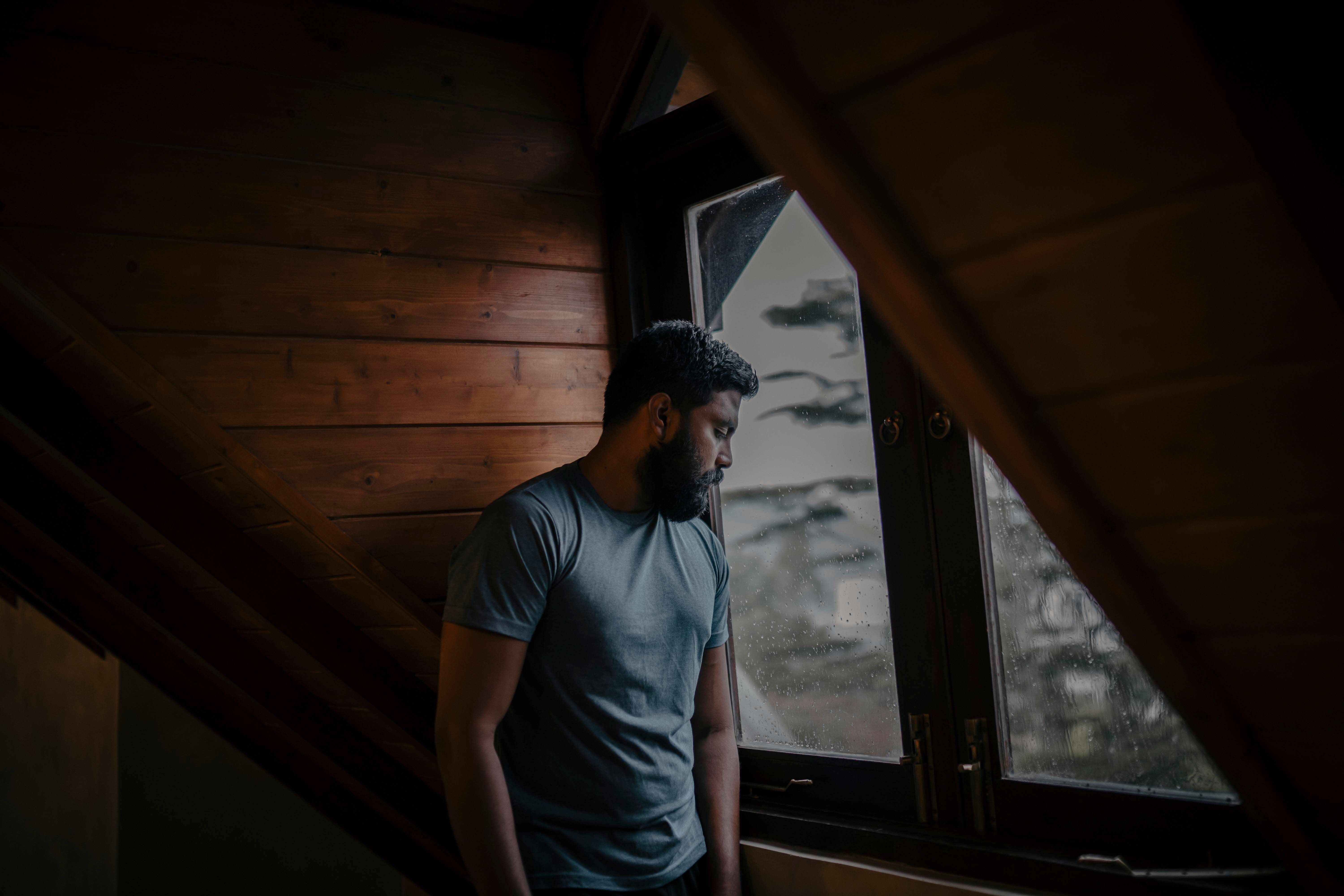Bearded man in a gray shirt standing in a dark wood paneled room with his eyes closed leaning against the wall beside a rain streaked window. Betrayal trauma doesn't just hurt, it lives in the body. Trauma therapy in Milwaukee, WI helps men release what they've been carrying and start healing from the inside out.