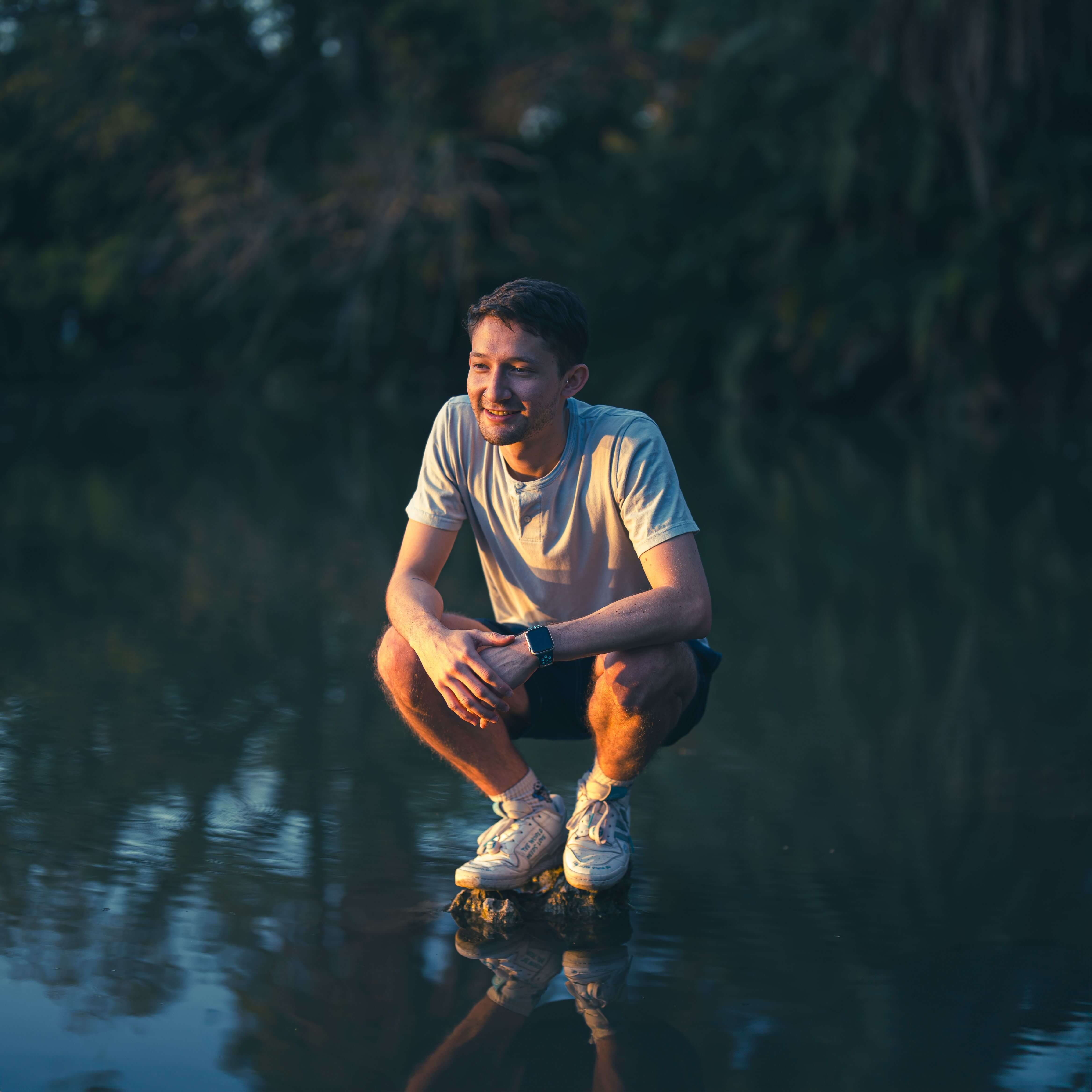 Young man in a white shirt crouching on a rock in shallow water smiling softly with trees and water reflections behind him in warm evening light. Healing from betrayal trauma is possible. Trauma therapy in Milwaukee, WI helps men rebuild trust in themselves and rediscover a sense of peace and purpose.