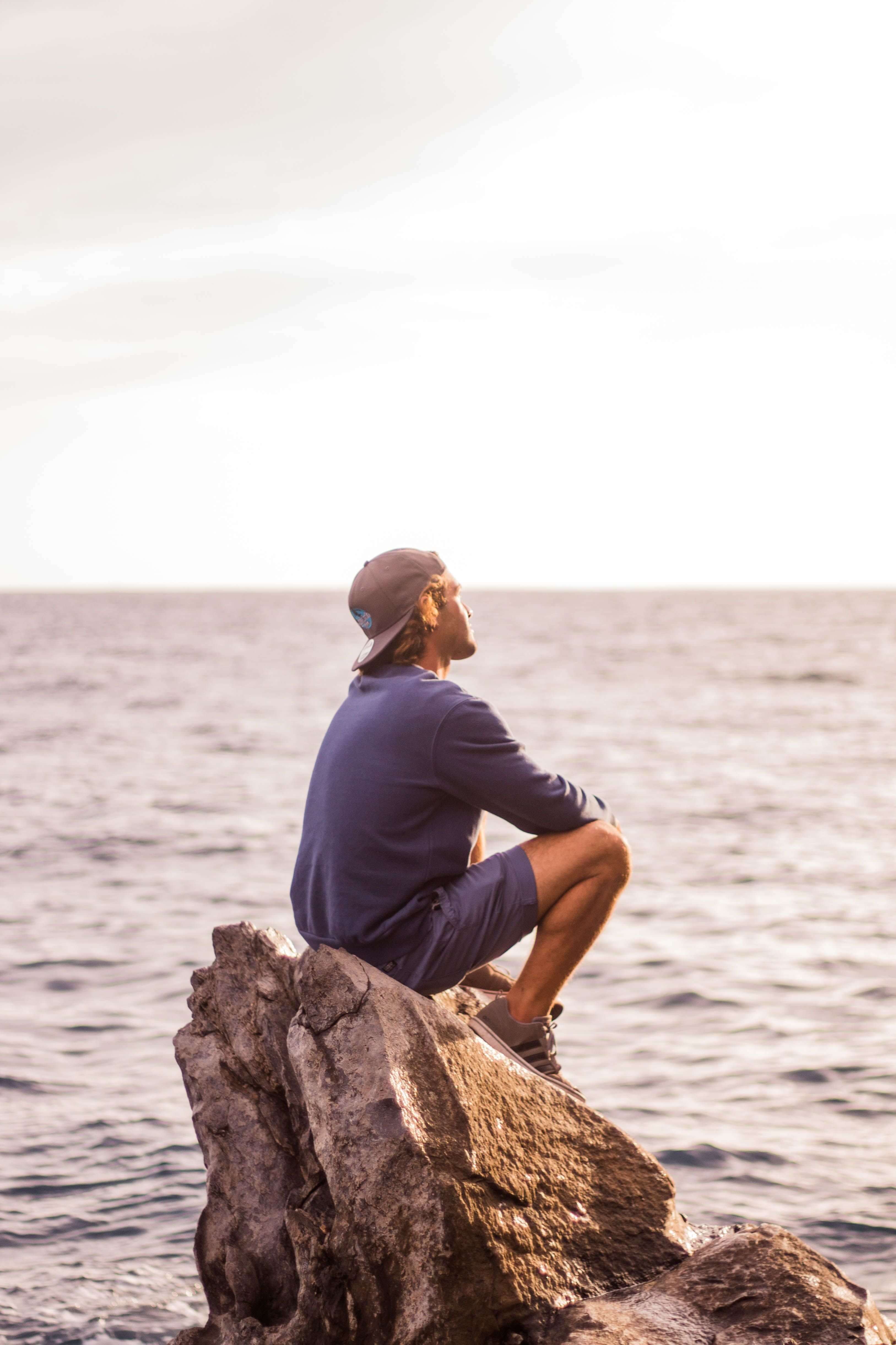 Man in a blue shirt and cap sitting on a rock at the ocean's edge looking upward toward a bright overcast sky. Healing is possible when you address trauma where it actually lives. Trauma therapy in Milwaukee, WI helps men regulate their nervous system and reclaim their life.