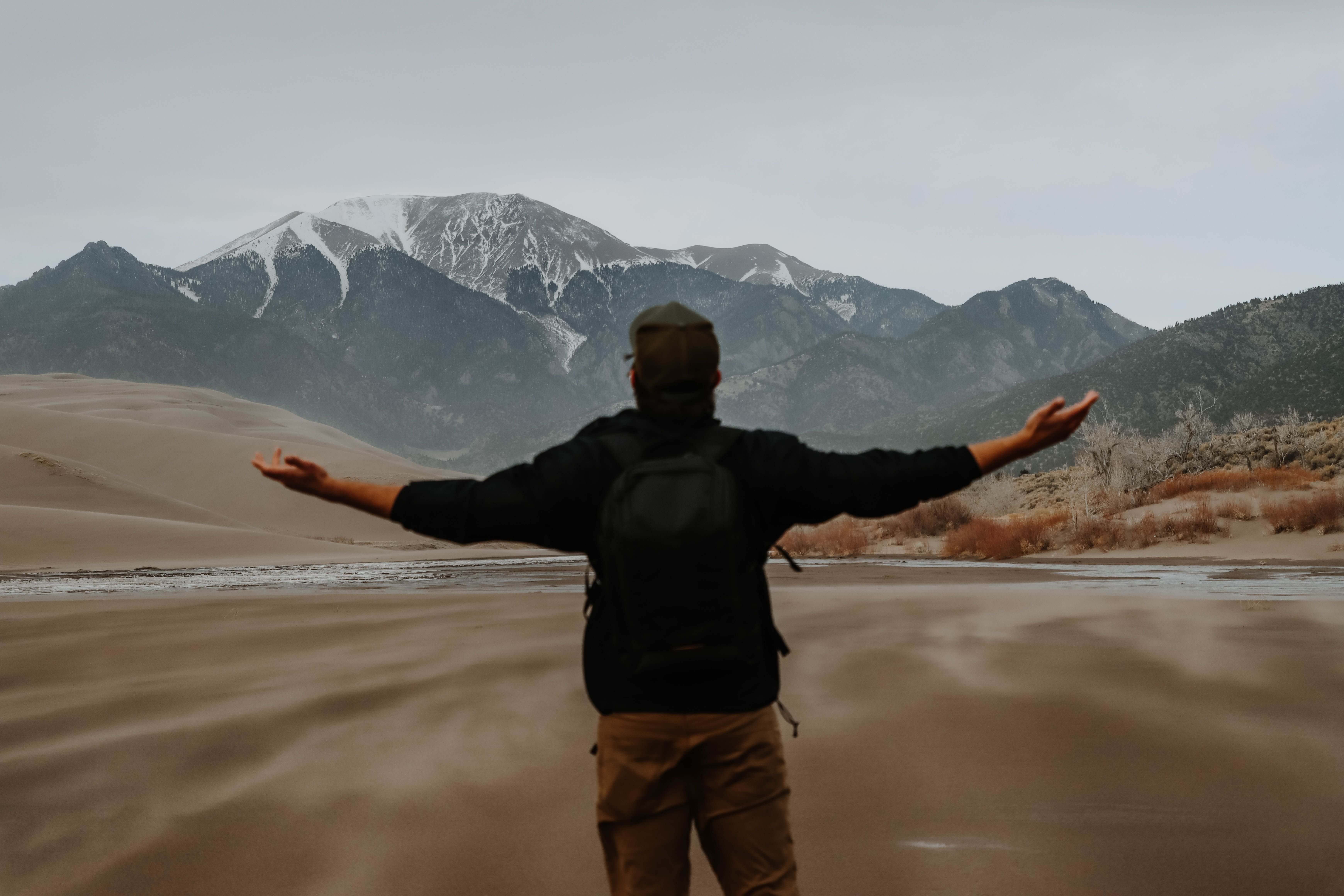 man with backpack arms | trauma therapy in Milwaukee, WI | how to heal from trauma | mens trauma therapy | Oak Creek | River Hills Man in a black jacket and backpack standing with both arms outstretched facing vast sand dunes and snow capped mountains under a cloudy sky. Trauma therapy in Milwaukee, WI helps men reconnect with purpose and feel like themselves again.