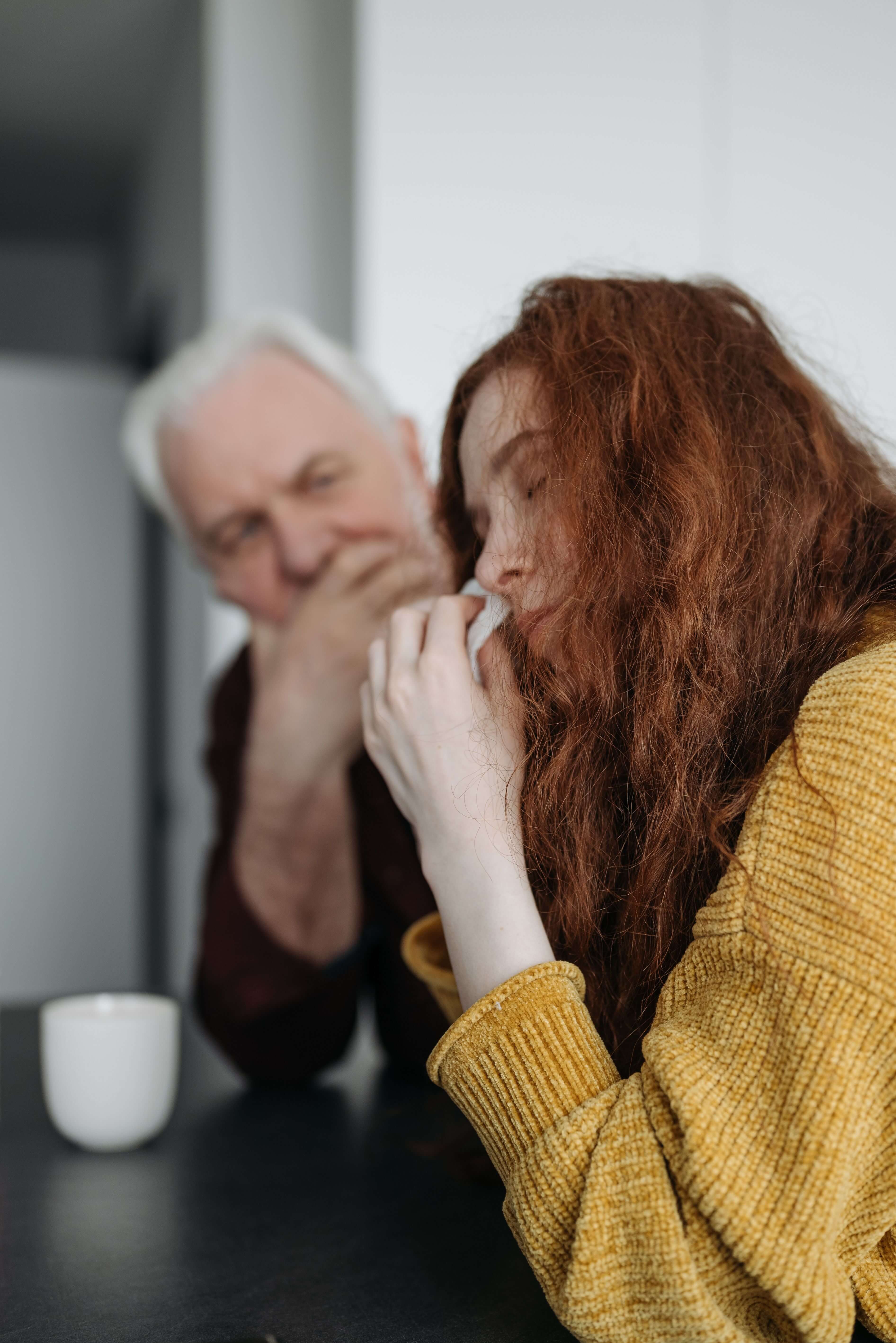 Woman with long red hair in a yellow sweater sitting at a table with her head down while an older man with white hair looks on with concern in the background. Trauma therapy in Milwaukee, WI helps men process vicarious trauma and reconnect with the people they care about.