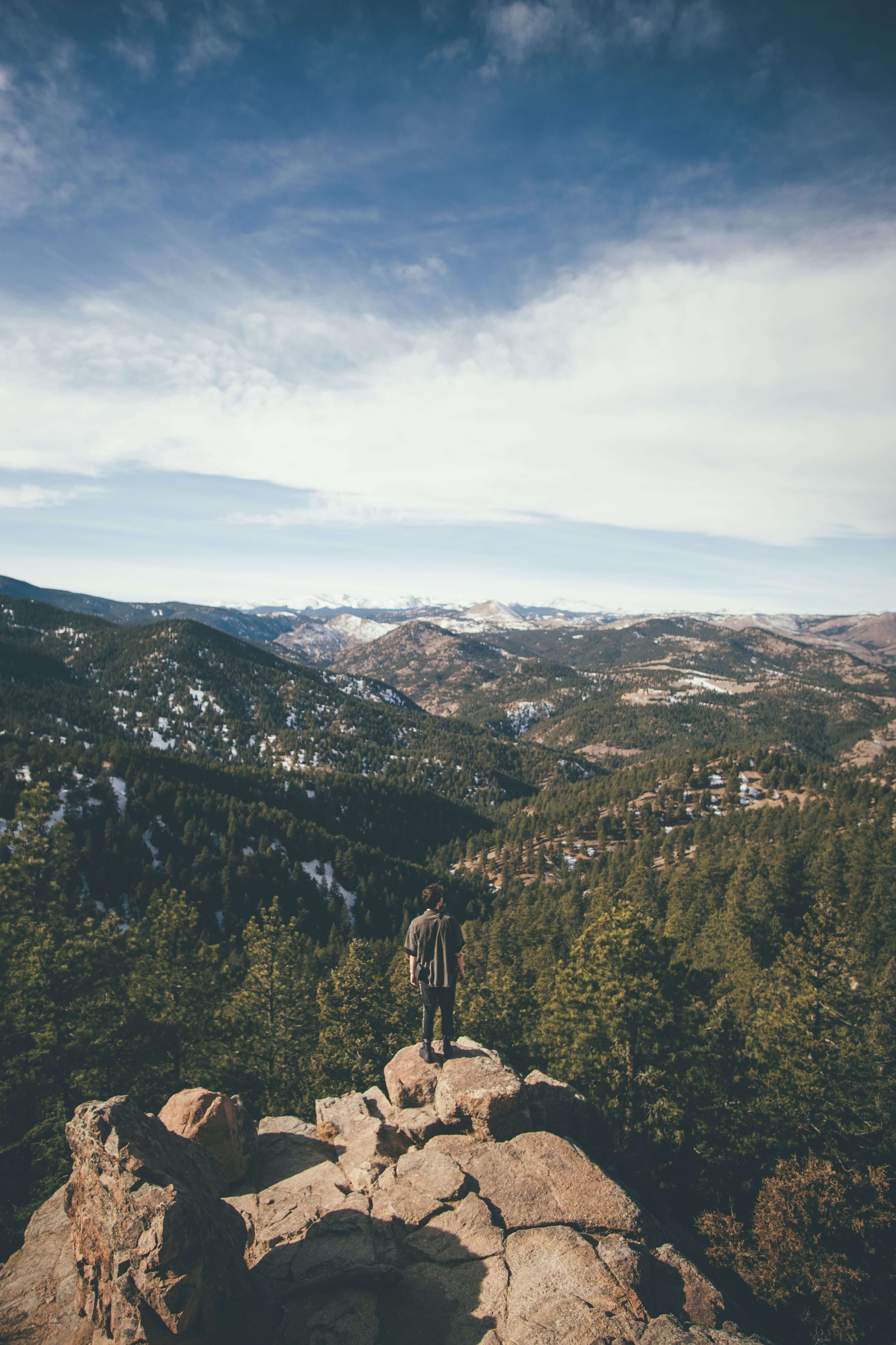 man standing alone on rocky overlook | trauma therapist in Milwaukee, WI | mens trauma therapy | how to heal from trauma | Wauwatosa | Brookfield | Pewaukee Small figure of a man standing alone on a rocky mountain outcrop overlooking a vast forested valley with snow capped peaks in the distance. Trauma therapy in Milwaukee, WI helps men find their way back to who they truly are.