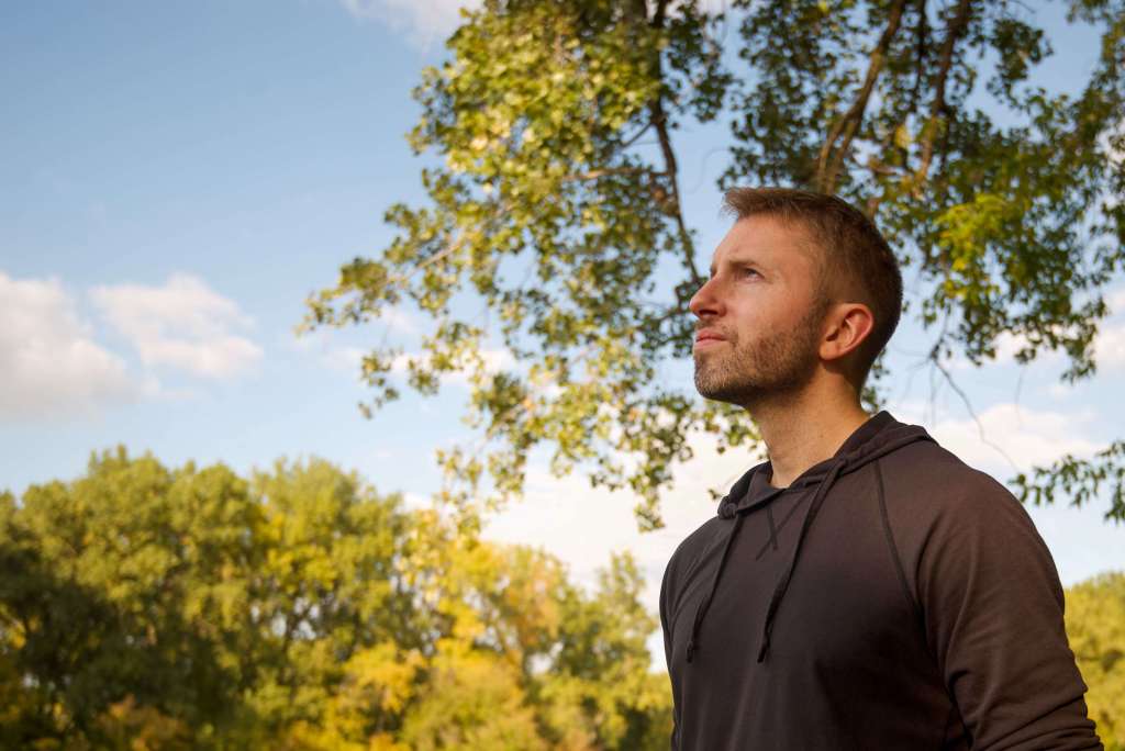 Man in a dark hoodie standing outdoors looking upward with trees and a blue sky behind him in warm natural light. When trauma lifts you can finally look up and take it all in. Trauma therapy in Milwaukee, WI helps men reconnect with presence and find their way back to life.
