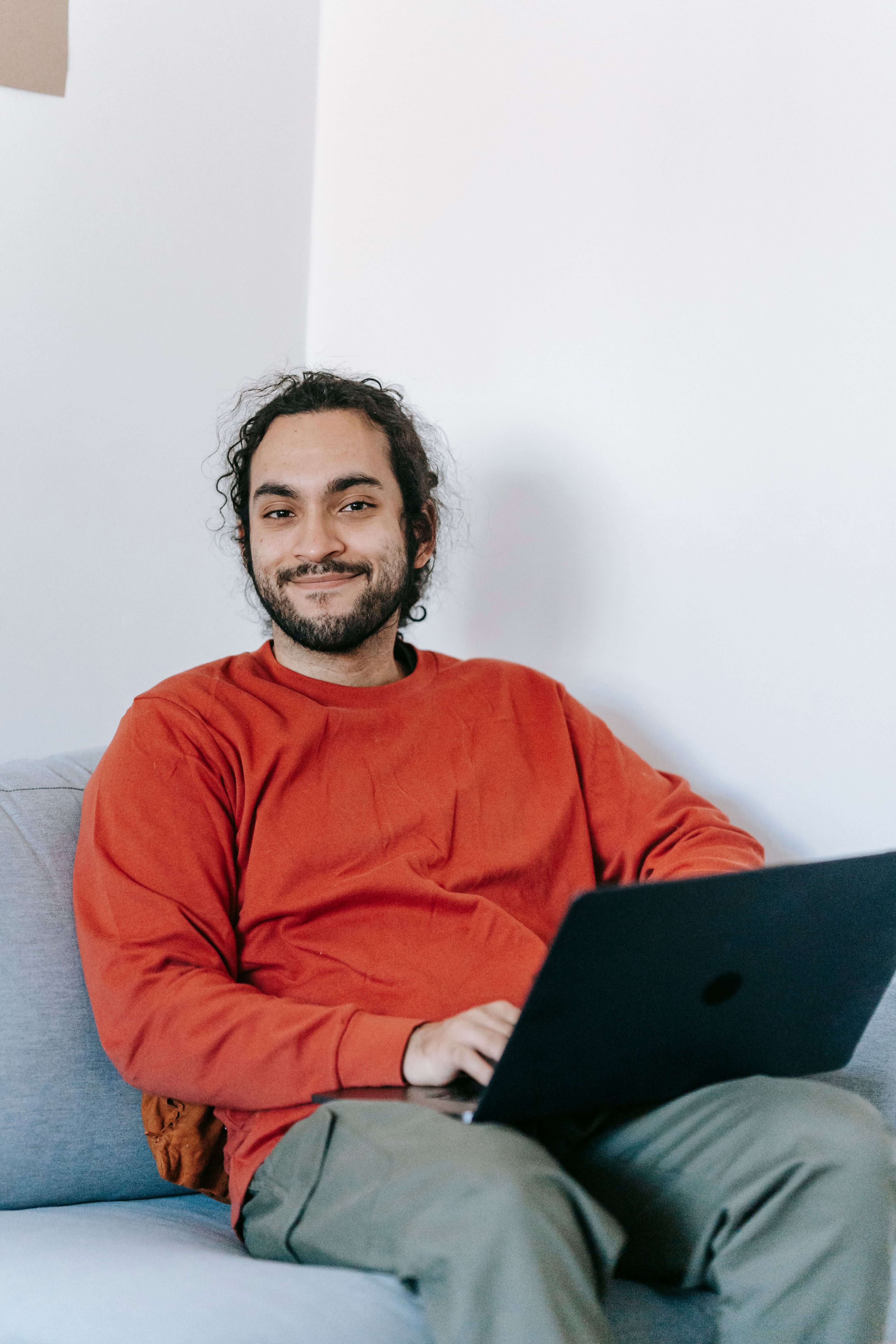 Young man with curly hair and a beard in a red sweater sitting on a light gray couch smiling while holding an open laptop. Virtual therapy is just as effective as in person. Online trauma therapy in Milwaukee, WI helps men start healing from wherever they feel most comfortable and safe.