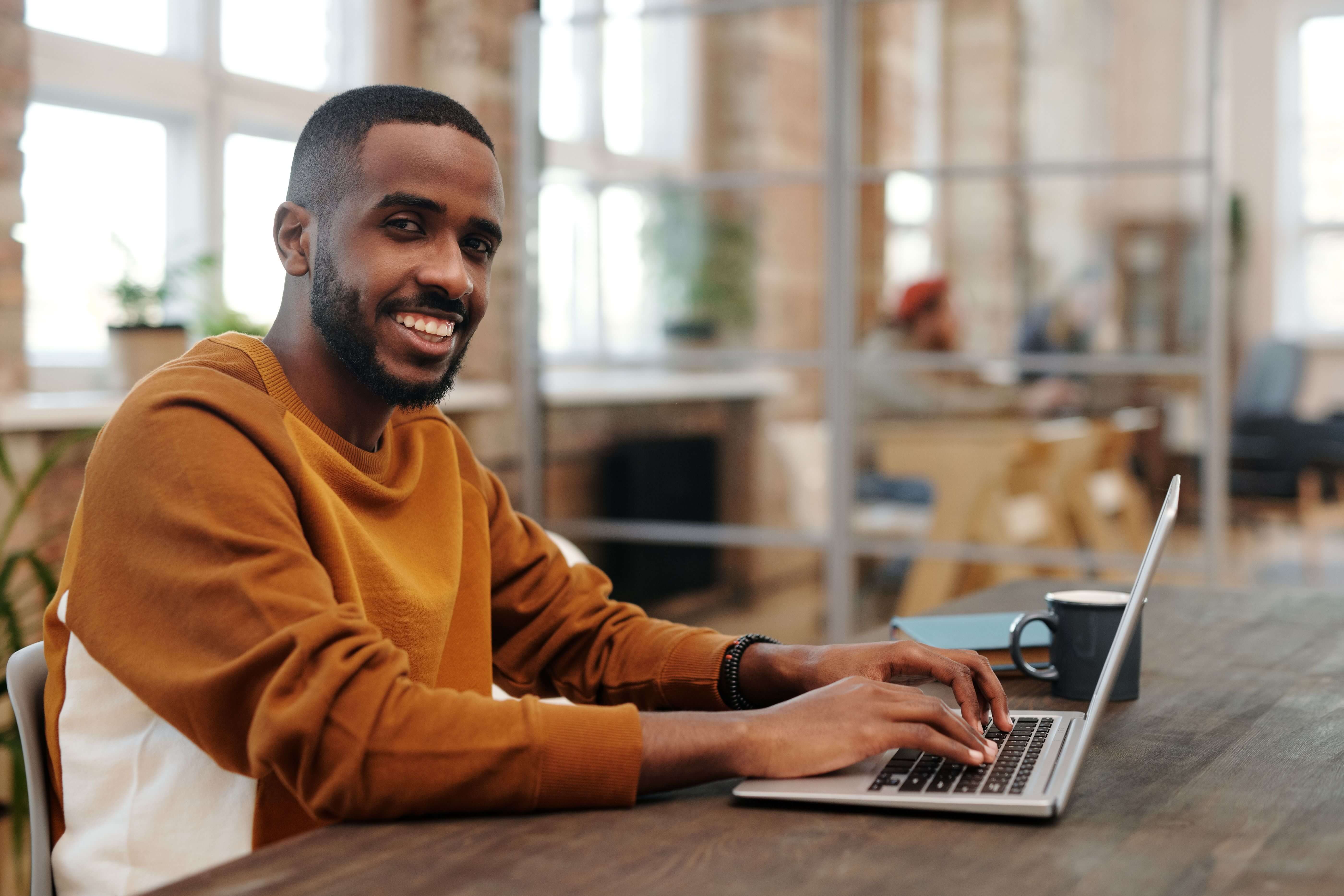 Smiling man in an orange sweater sitting at a wooden desk typing on a laptop in a bright open workspace with brick walls and plants behind him. Healing is possible from wherever you are. Online trauma therapy in Milwaukee, WI gives men access to quality care without leaving the comfort of home.