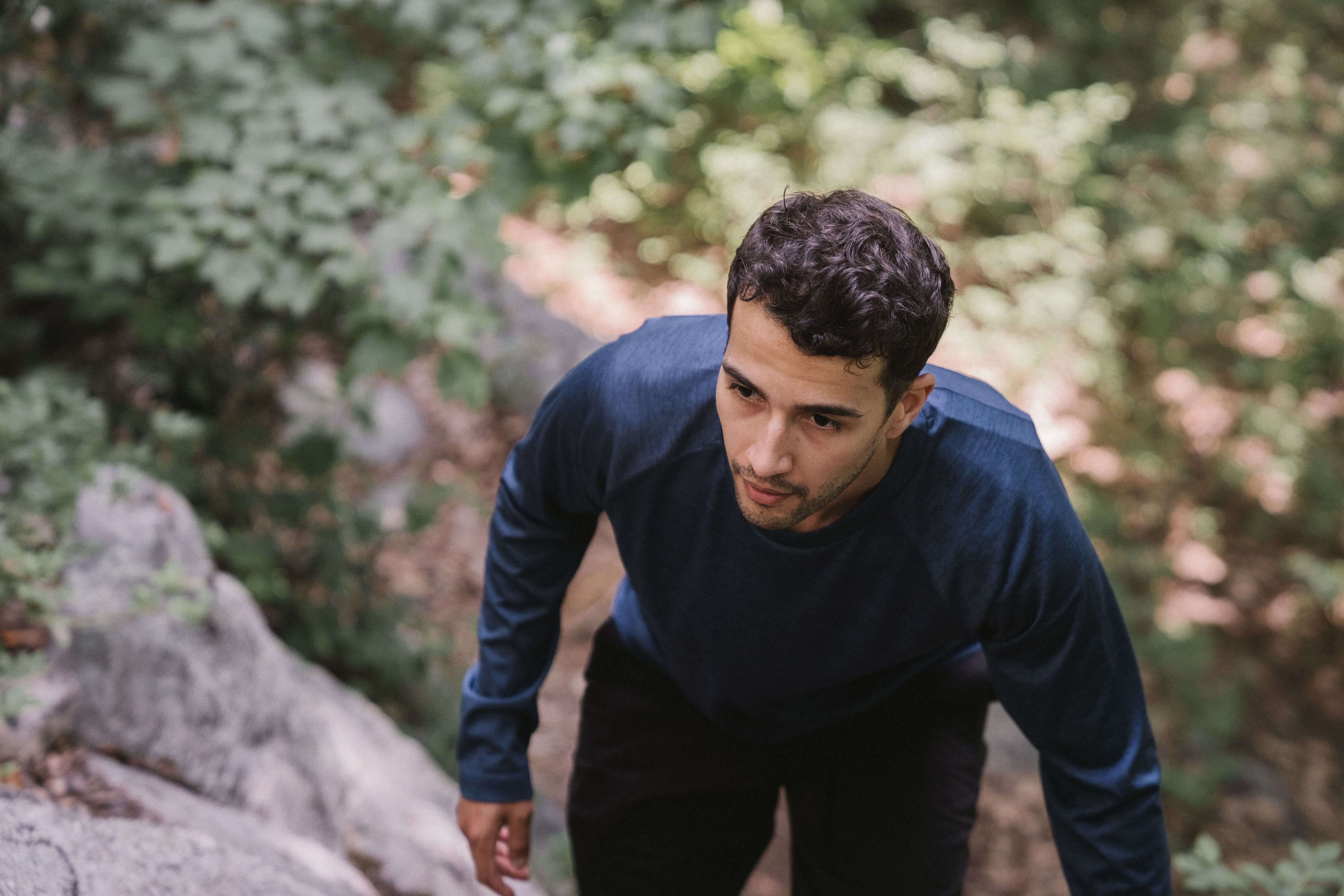 young man in navy shirt | mens trauma therapy in Milwaukee, WI | trauma in men | therapy for men feeling disconnected | Oak Creek | River Hills Young man with curly hair in a navy long sleeve shirt climbing up a rocky trail through a green wooded area with a focused expression. Healing from disconnection takes courage and one step at a time. Trauma therapy in Milwaukee, WI helps men climb out of survival mode and back into their own life.
