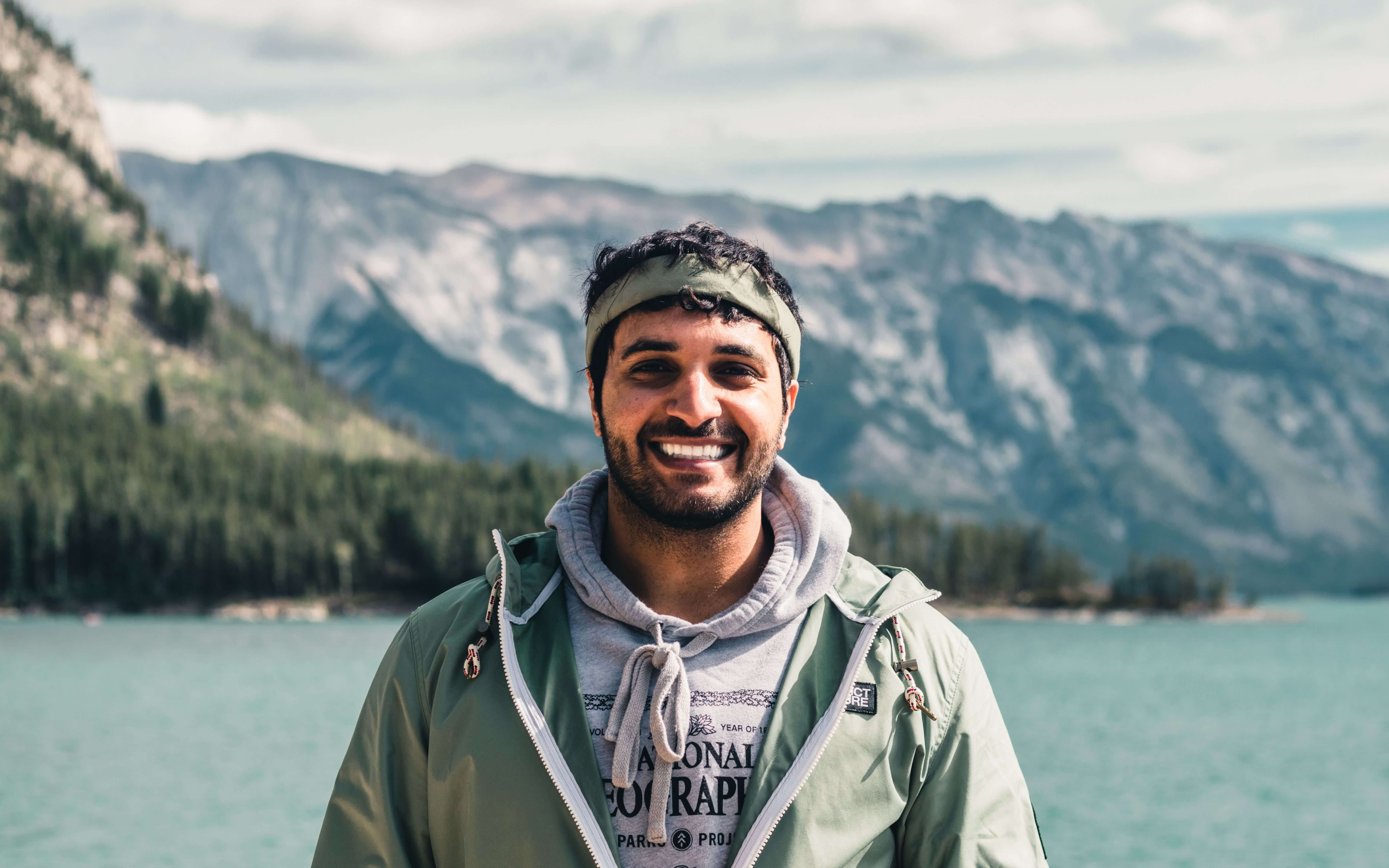 Smiling young man with a headband and green jacket standing outdoors with a turquoise mountain lake and rocky peaks behind him. Trauma therapy in Milwaukee, WI helps men rebuild trust and experience relationships that feel safe and real.