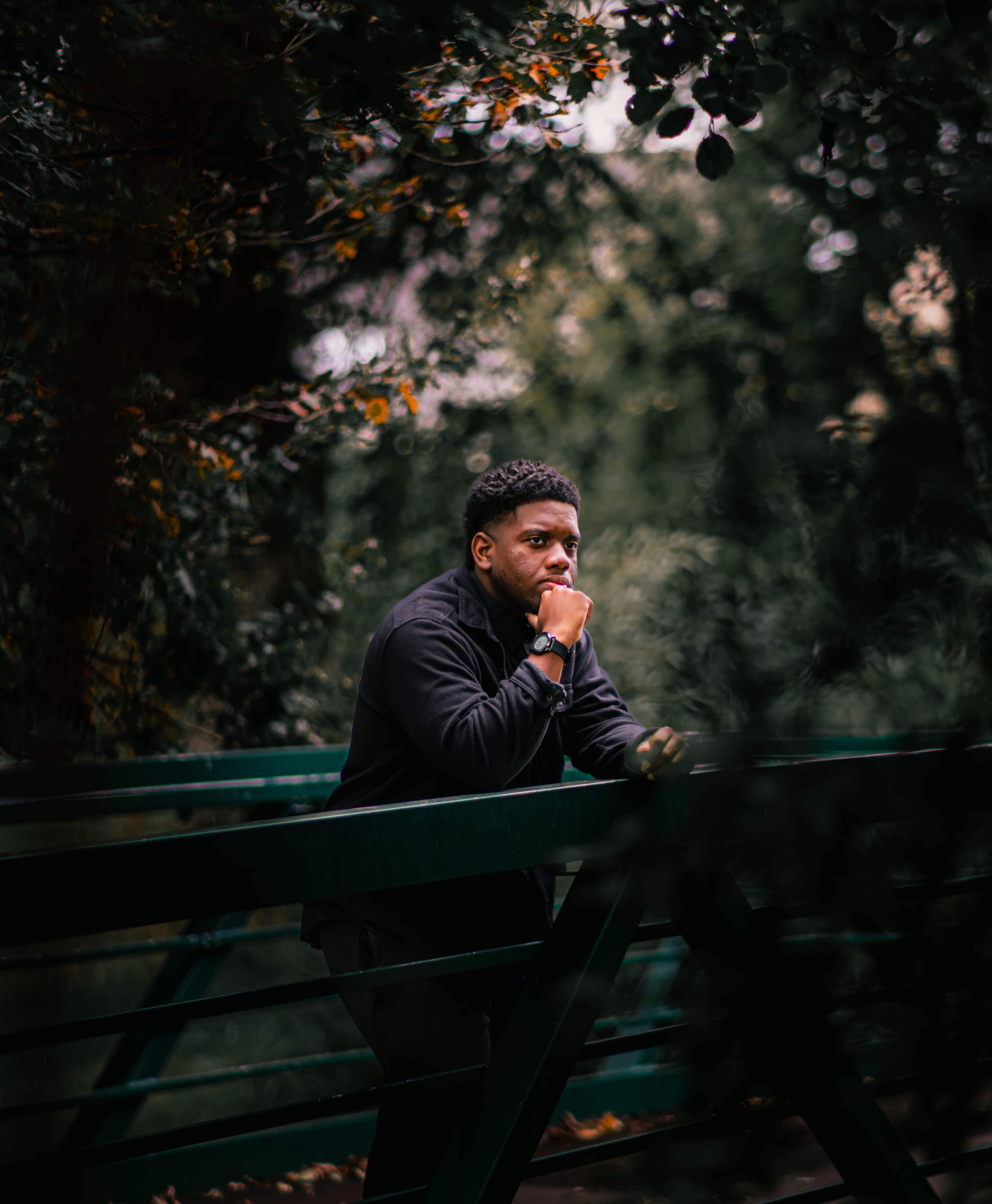 Young man in a dark jacket leaning on a green metal bridge railing with his chin resting on his hand surrounded by dark trees. Trauma therapy in Milwaukee, WI helps men understand why trust feels impossible and begin healing the relational wounds.