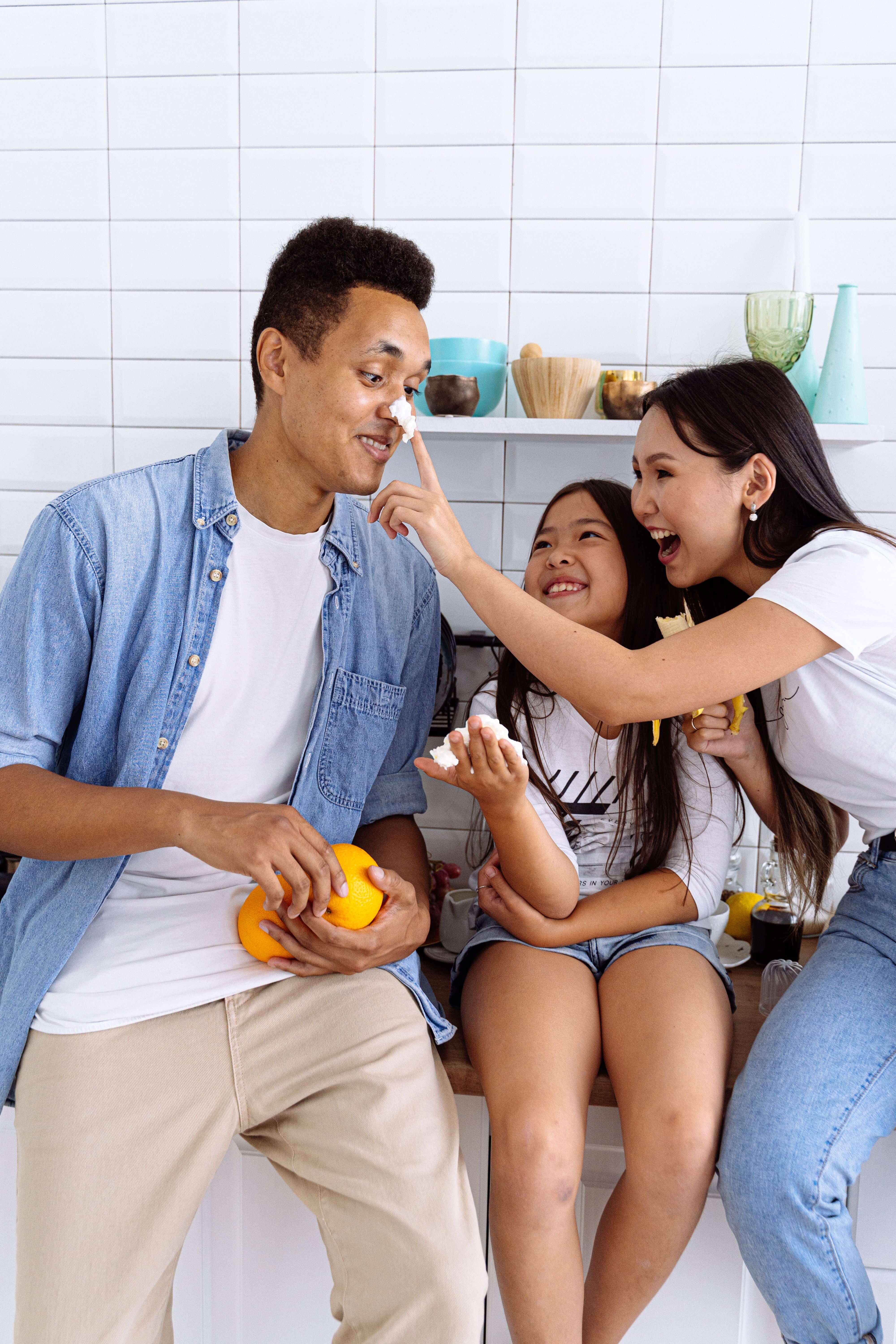man laughing with daughter and partner | trauma therapist in Milwaukee, WI | how to heal from trauma | mens trauma therapy | Pewaukee | Greenfield | Greendale Man in a blue denim shirt laughing with his daughter and partner in a bright kitchen while holding an orange. Healing from trauma means being fully present for the people you love. Trauma therapy in Milwaukee, WI helps men release stored physical symptoms and reconnect with their family.