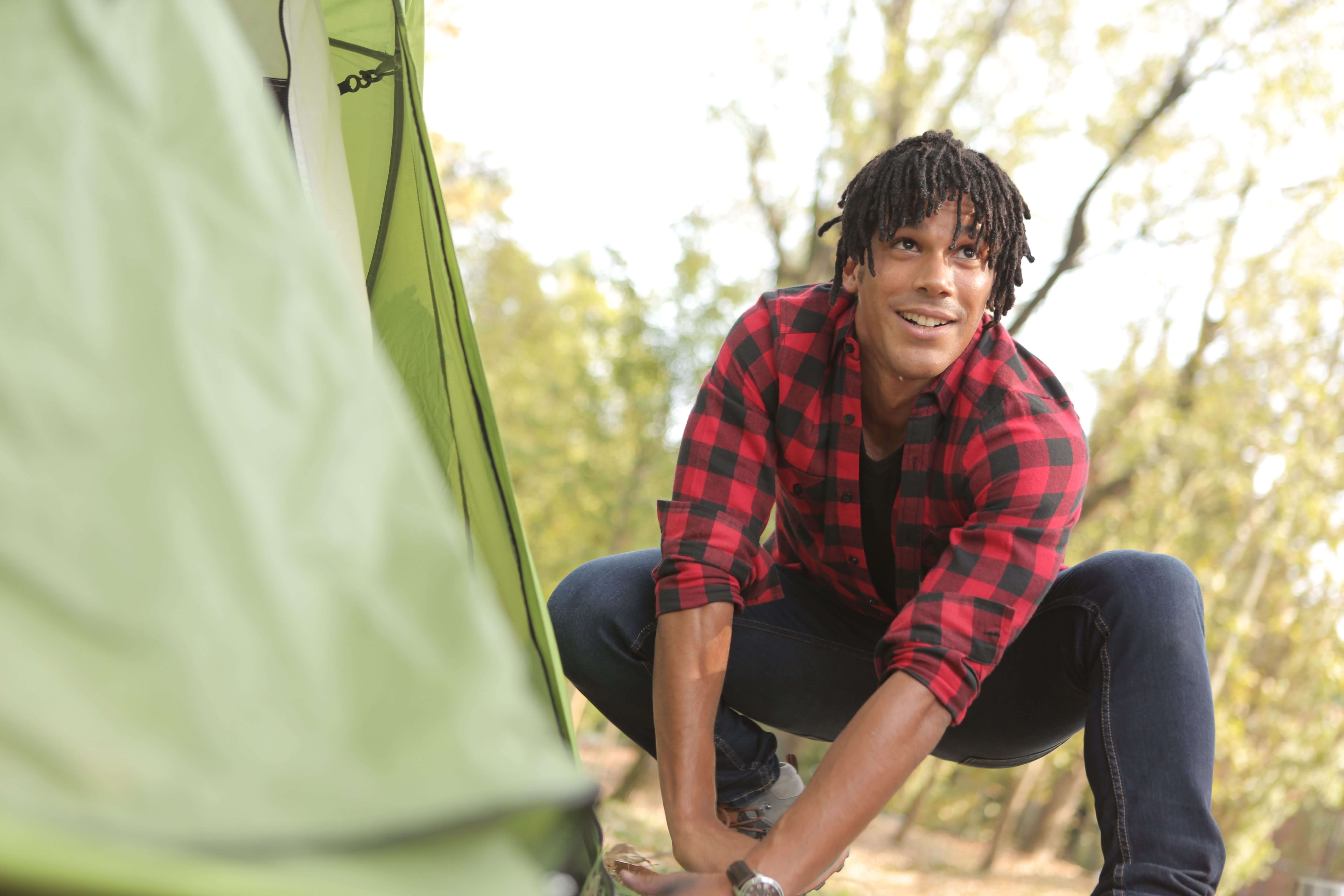 man with locs in plaid | mens trauma therapy | trauma symptoms | online trauma therapy | Oak Creek | River Hills Man with locs in a red and black plaid shirt crouching and smiling near a green tent in a wooded outdoor setting. Trauma therapy in Milwaukee, WI helps men heal from little t traumas and big T traumas alike so they can show up fully for the moments that matter.